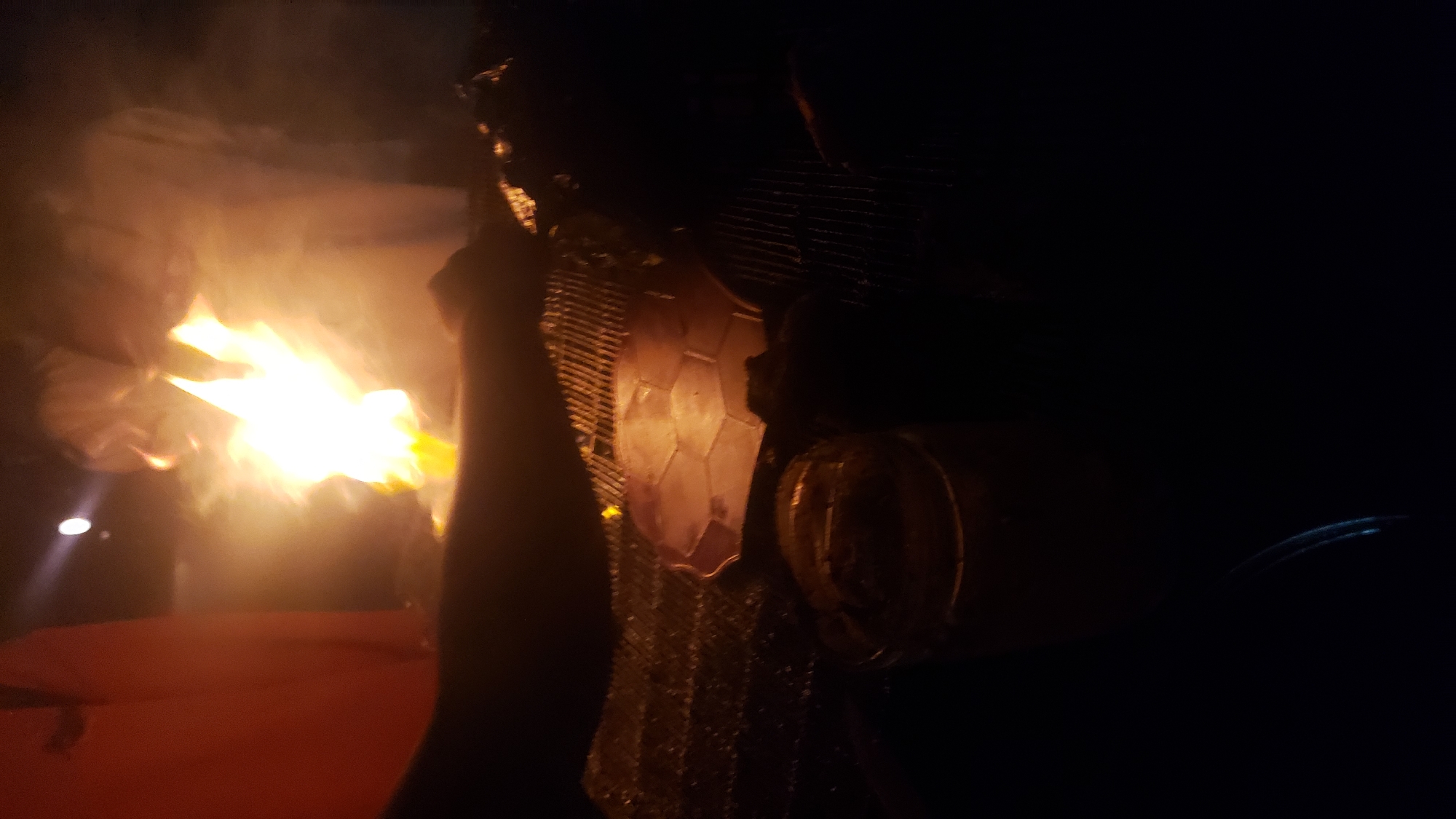 This image depicts a nighttime outdoor scene in Narayi, Nigeria, centered around a large, bright open fire. The fire, characterized by intense yellow and white flames, emits significant light and smoke, acting as the primary illuminant for the immediate area.
Behind the fire, two human figures are visible through the smoke and glare. They appear to be wearing light-colored clothing and are standing or bending, engaged in activity near the flames, consistent with tending to the fire or food. Their facial features and precise actions are obscured by the fire's intensity and smoke.
In the mid-ground, positioned over the fire, is a metal grill grate. On this grate, to the left of the fire's core, a light-colored, polygonal object with a distinct hexagonal pattern is present, likely a piece of food being cooked. Further to the right on the grill, darker, less defined items are visible, also undergoing preparation. A dark-skinned arm or limb extends from the left foreground towards the grill, suggesting a person is actively interacting with the cooking process.
In the lower left foreground, a clear glass or plastic container, possibly a jar, is partially visible. It contains a dark, granular substance and appears to be a drinking vessel or holding a condiment. The surrounding environment is predominantly dark, indicating a nighttime setting, with shadows cast by the fire's strong light. A faint, distant light source is visible in the upper left background.