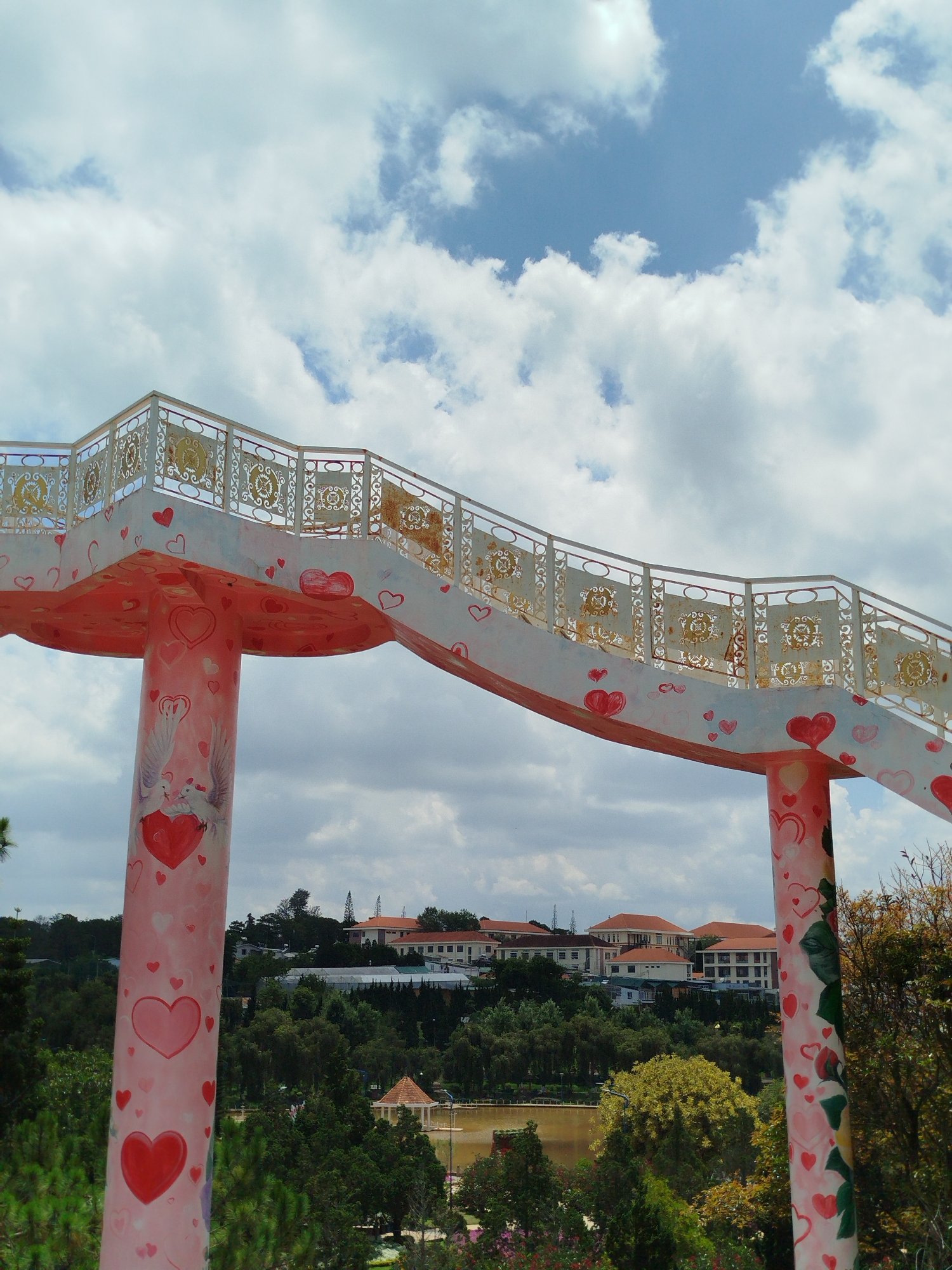 The image captures a vibrant, decorated pedestrian bridge curving across the upper frame, set against a bright, cloudy sky with patches of blue. The bridge's main structure is painted in shades of pink and white, lavishly adorned with numerous red and pink hearts of various sizes. One of the prominent support pillars, seen on the left, features a detailed mural of two white doves with outstretched wings, facing each other over a large red heart, also surrounded by smaller hearts. The bridge's railings are white, featuring intricate golden-hued scrollwork patterns, some of which exhibit signs of wear or rust.
Below the bridge, a lush landscape unfolds, dominated by a dense canopy of green trees, including some with lighter, almost golden-green foliage. In the mid-ground, a calm body of water, possibly a lake or pond, is visible, with a small gazebo-like structure on its edge. Further in the background, nestled among the trees on a gentle hillside, are several buildings with distinctive red-tiled roofs, typical of architecture found in Da Lat, Vietnam. The scene is brightly lit, indicating daytime under a partly cloudy sky. No people or specific activities are visible, but the heart and dove motifs on the bridge evoke themes of love, romance, and peace, suggesting it may be a popular spot for visitors. There is no visible text in the image.