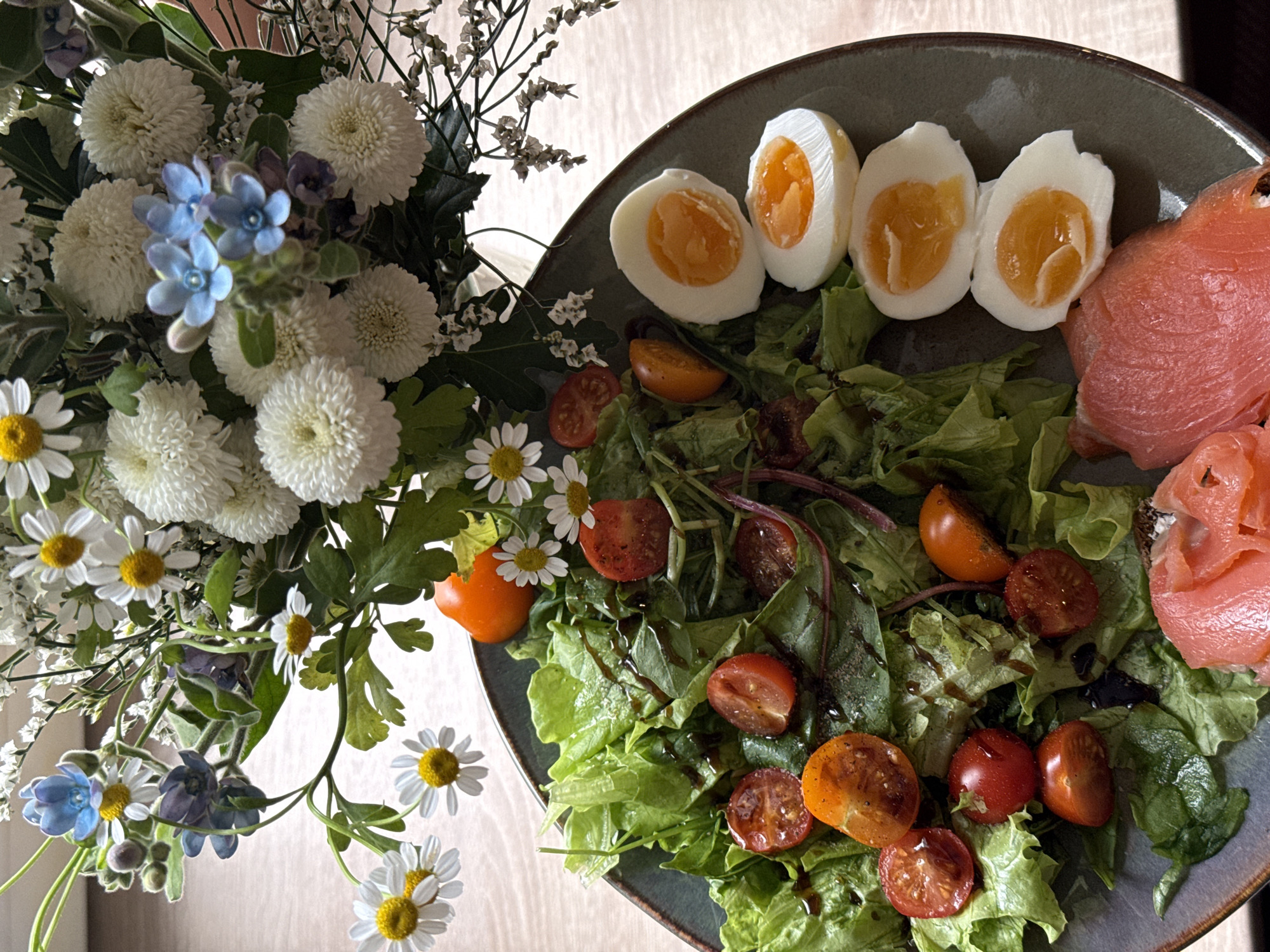 The image features a plate with a salad, hard-boiled eggs, and smoked salmon alongside a bouquet of flowers. The salad consists of mixed greens and halved cherry tomatoes, drizzled with a dark dressing. Three halved hard-boiled eggs are positioned on the plate, with the yolks still soft. Slices of smoked salmon are adjacent to the eggs. The flowers, arranged in a bouquet, include white chrysanthemums, blue forget-me-nots, and white daisies. The scene is set on a light-colored wooden table. The image provides no specific details that would indicate its location in Krasnoyarsk, Russia, or any other city.