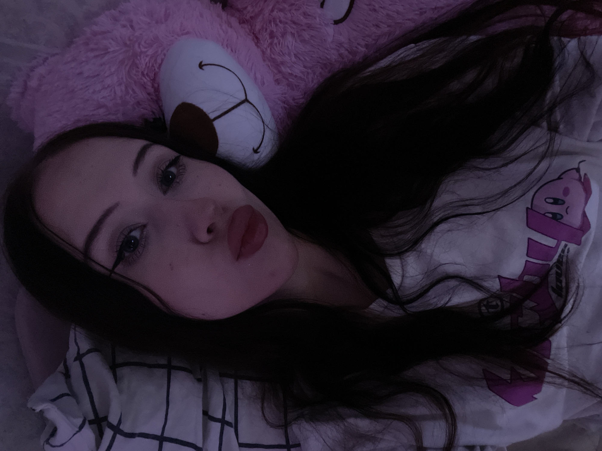 The media file shows a young person with long, dark hair lying down, possibly in a bed. They are positioned close to a large, plush pink teddy bear, which is mostly out of focus but its white face with brown ears and embroidered features is visible. The person is wearing a white t-shirt with a pink character design on it, possibly an anime character. The background includes a pillow with a black and white checkered pattern.
The lighting in the image is dim, suggesting it might be nighttime or a dimly lit room. The overall mood appears relaxed and comfortable, with the person's eyes open and looking towards the camera. The close-up perspective emphasizes the person's facial features, including their eyes, eyebrows, and lips, which appear to be lightly made up. A single strand of hair falls across the person's face, obscuring part of their eye.
There is no visible text in the image. The setting appears to be indoors, likely a bedroom, given the presence of a bed, pillow, and plush toy. The absence of any outdoor elements or specific interior decorations makes it difficult to ascertain a more precise location beyond being indoors. The location context provided is Votkinsk, Russia, but there are no visual cues in the image to confirm this.