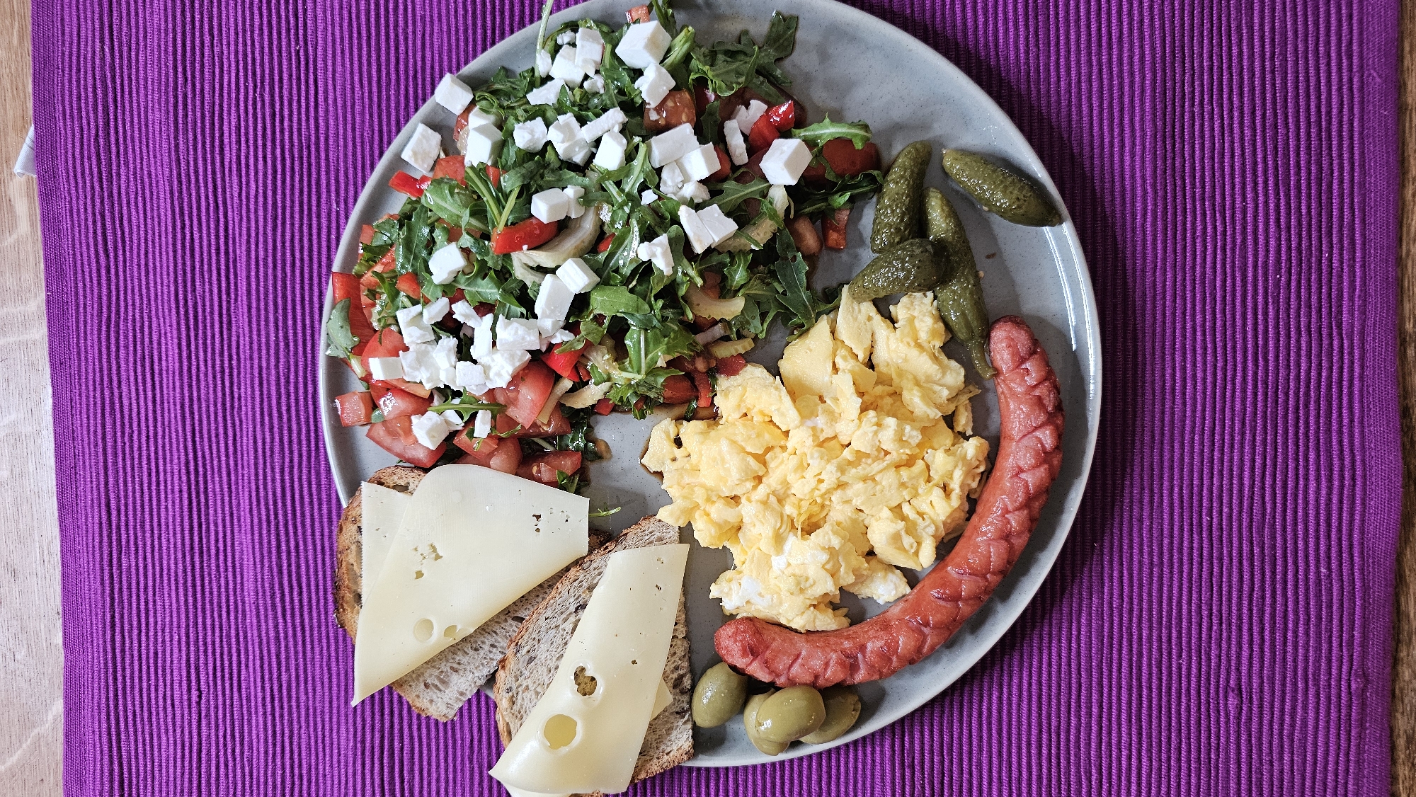 The image presents an overhead view of a varied meal served on a round, light grey plate. The plate is positioned on a vibrant purple, ribbed placemat, which in turn rests on a light-colored wooden surface.
The meal consists of several components:
In the upper left, a fresh salad features green arugula, diced red tomatoes, and possibly red onion or bell pepper, all generously topped with crumbled white cheese, likely feta.
Adjacent to the salad, in the center of the plate, is a serving of fluffy scrambled eggs.
To the right of the eggs, a single, reddish-brown sausage with a textured surface is placed.
Above the sausage, several small, green gherkins or pickled cucumbers are visible.
In the lower left, two slices of dark, possibly whole-grain bread are topped with slices of pale yellow cheese, one showing distinct holes.
A small cluster of green olives completes the food arrangement on the plate.
The scene suggests an indoor setting, likely a dining area, with the meal prepared and ready to be eaten, resembling a breakfast or brunch spread. There are no people visible, and no text is present in the image.