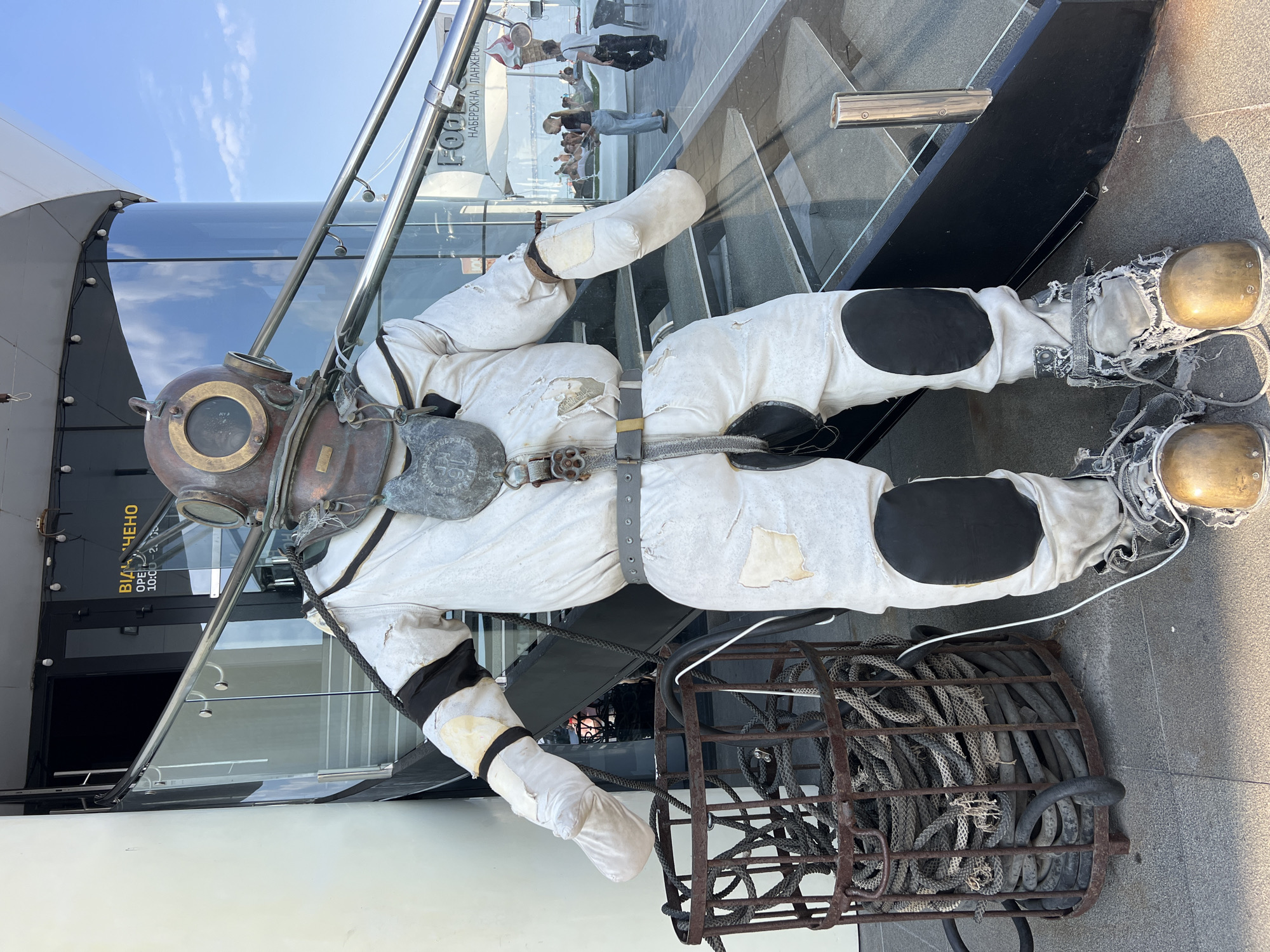 A full-body, vintage diving suit mannequin stands outside on a sunny day. The suit is off-white with dark patches on the knees and elbows, and appears to be made of heavy fabric. It has a large, metal helmet with round glass windows. A thick rope is coiled inside a rusted metal basket next to the mannequin. The mannequin is positioned on a tiled ground in front of a modern building with large glass windows and a metal staircase. In the background, people can be seen walking and sitting on a terrace overlooking the sea. The location is Odesa, Ukraine, specifically on the Odesa embankment, near the "Food" cafe and ice cream stand. The building appears to be a part of the Odesa Sea Port or a waterfront establishment. The sky is clear blue with a few white clouds.