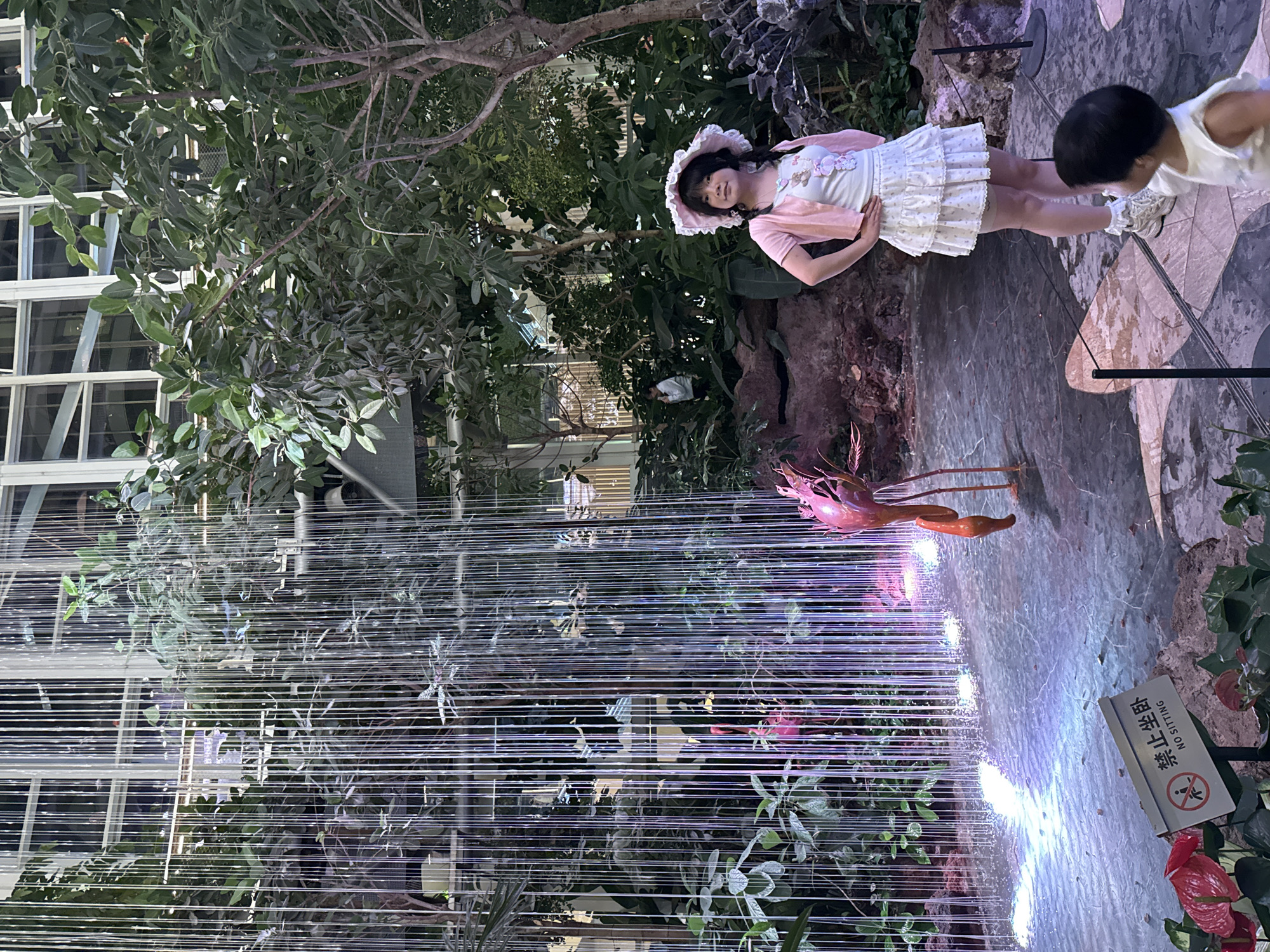 In Liangjiang, China, a young woman dressed in a pink cardigan and white ruffled skirt stands with her hands on her hips in front of a curtain of lights that simulates a waterfall. She wears a wide-brimmed hat and has her hair in braids. To her left, a decorative flamingo stands near the illuminated water feature. A small child in a white top is visible in the foreground, looking away from the camera. The scene appears to be an indoor botanical garden or a themed attraction, with lush greenery and modern lighting. A sign that reads "NO SITTING" in both Chinese and English is positioned near the water's edge.
