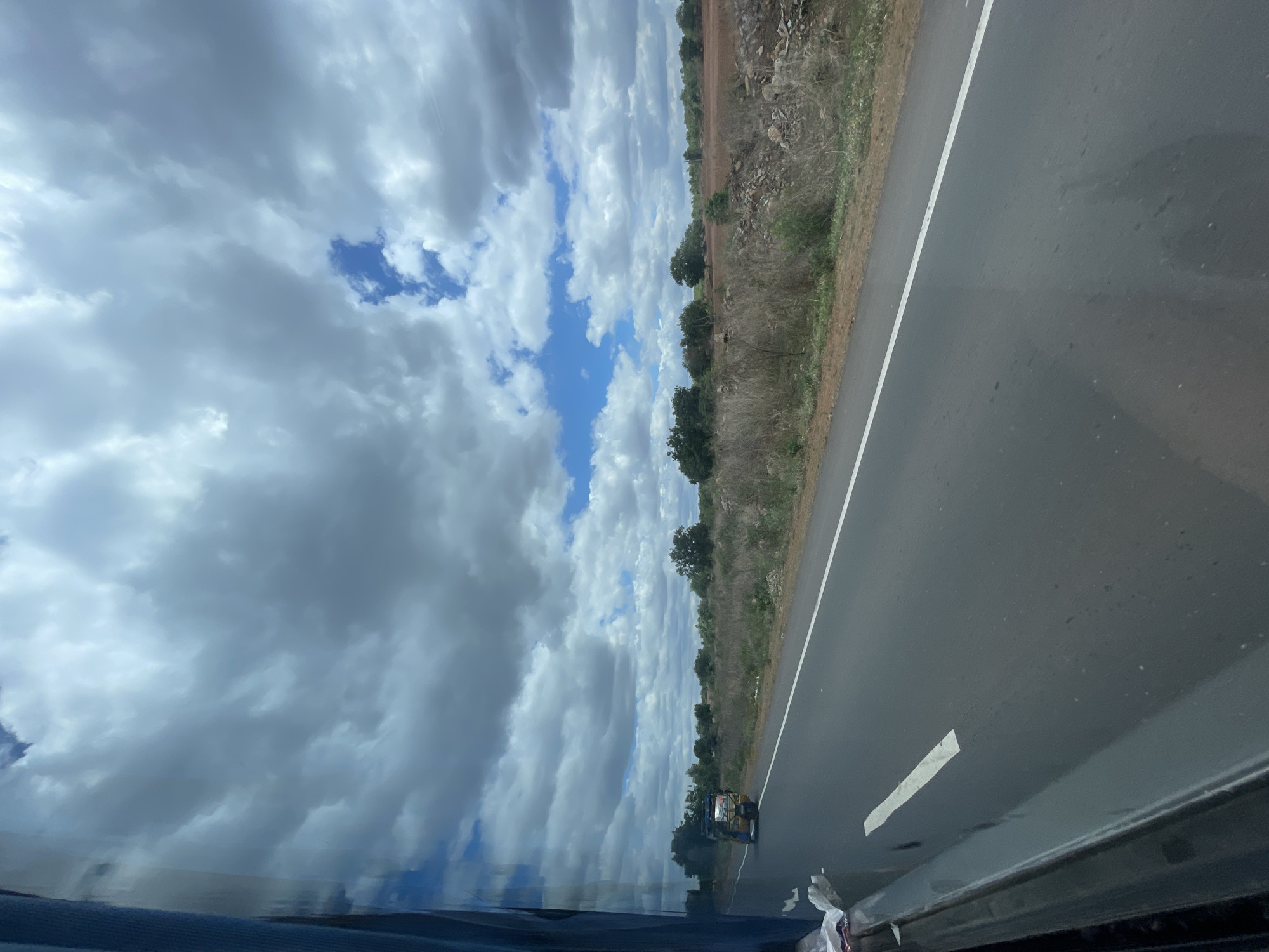 A view from inside a vehicle looking out at a two-lane road in Sugali Tanda, India. The road is paved and has a white line marking the lane. A yellow and blue autorickshaw is traveling on the road in the distance. The landscape beside the road is dry with sparse vegetation and scattered rocks. In the background, there are trees and a red, rocky field. The sky is filled with dramatic, gray and white clouds, with patches of blue sky visible. The reflection of the vehicle's interior is visible on the left side of the image.
