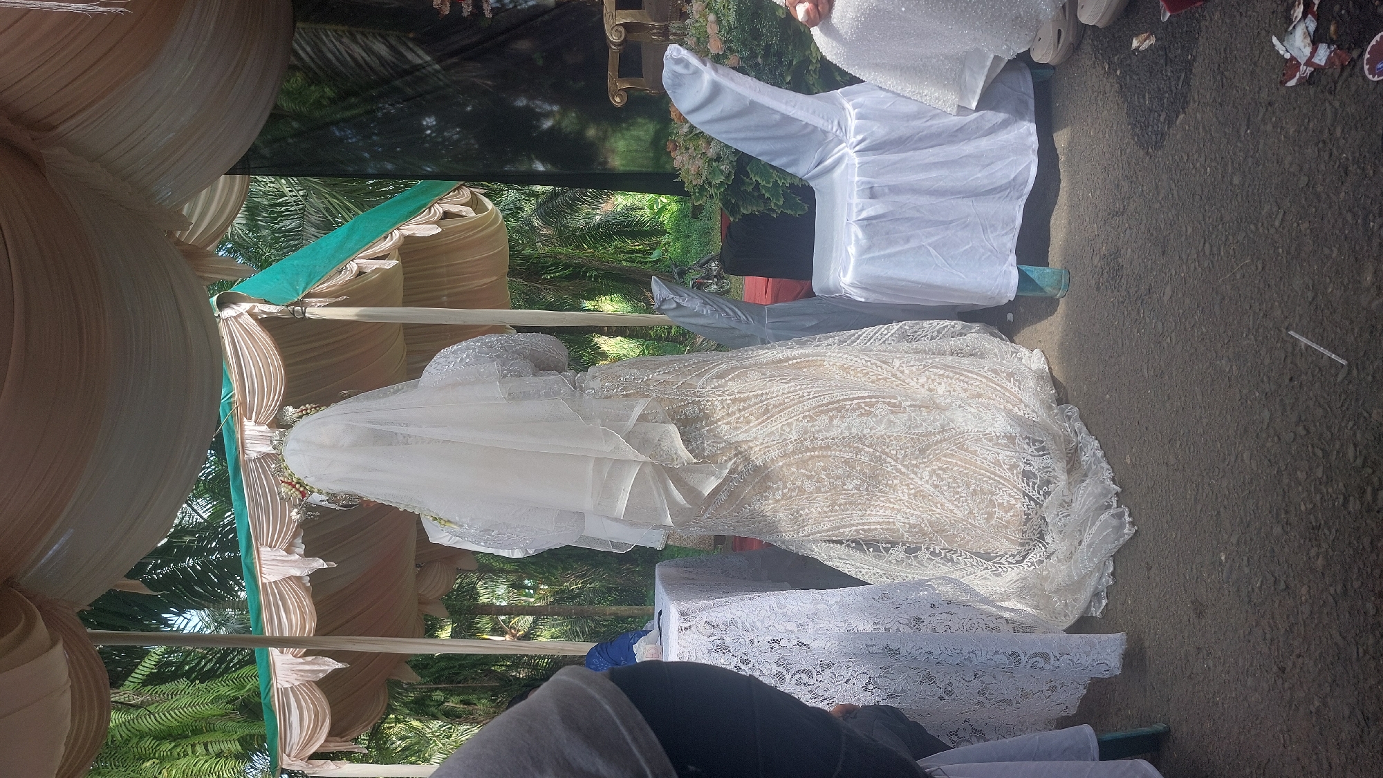 The image depicts an outdoor wedding ceremony in Sigara Gara, Indonesia, under a temporary canopy with beige and green fabric draping. A bride, facing away from the camera, is the central subject. She wears an elaborate white wedding gown with intricate lace detailing and a long veil. To her left, a table covered with a white lace tablecloth is partially visible. To her right, a white-covered chair is positioned. In the background, palm trees and lush greenery indicate a tropical environment. The ground appears to be paved with asphalt. The scene suggests a traditional Indonesian wedding ceremony.