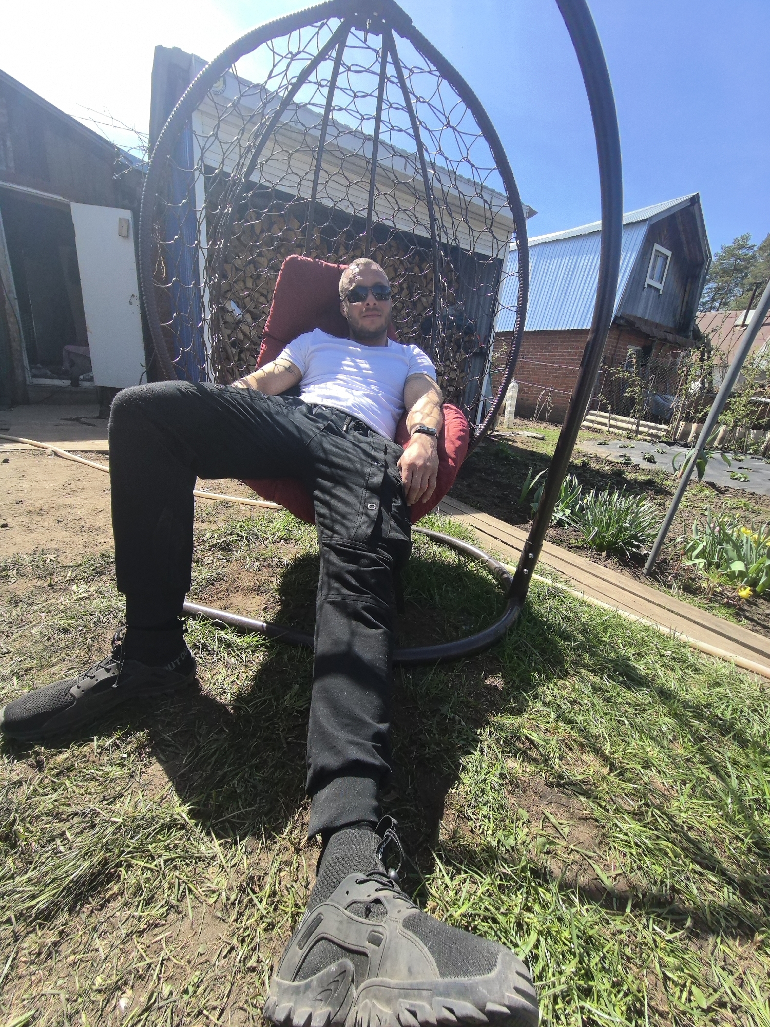 A man is seated in a hanging chair in a grassy yard. He wears black pants, a white t-shirt, sunglasses, and black shoes. The chair is reddish and made of woven material. Behind the chair is a shed with an open white door. To the right is a two-story building with a silver metal roof and a window. The ground is a mix of grass and bare dirt. It is daytime. The location cannot be verified.