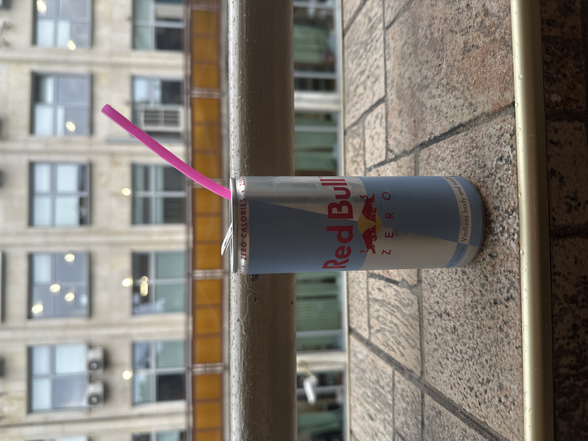 A light blue and white Red Bull Zero energy drink can, featuring a prominent red bull logo and "ZERO" text, rests on a wet, tiled outdoor surface. An upright pink straw extends from the opened can, which shows water droplets on its exterior. A light-colored, painted railing, also covered in water droplets and exhibiting minor paint chipping, is positioned horizontally in the mid-ground. The background reveals a blurred, multi-story building with a light-colored facade, numerous rectangular windows reflecting interior lights, and a horizontal architectural band of orange panels. Two external air conditioning units are visible on the building's exterior. The scene is illuminated by diffused, overcast light. The location is specified as Stara Zagora, Bulgaria. No individuals are present.