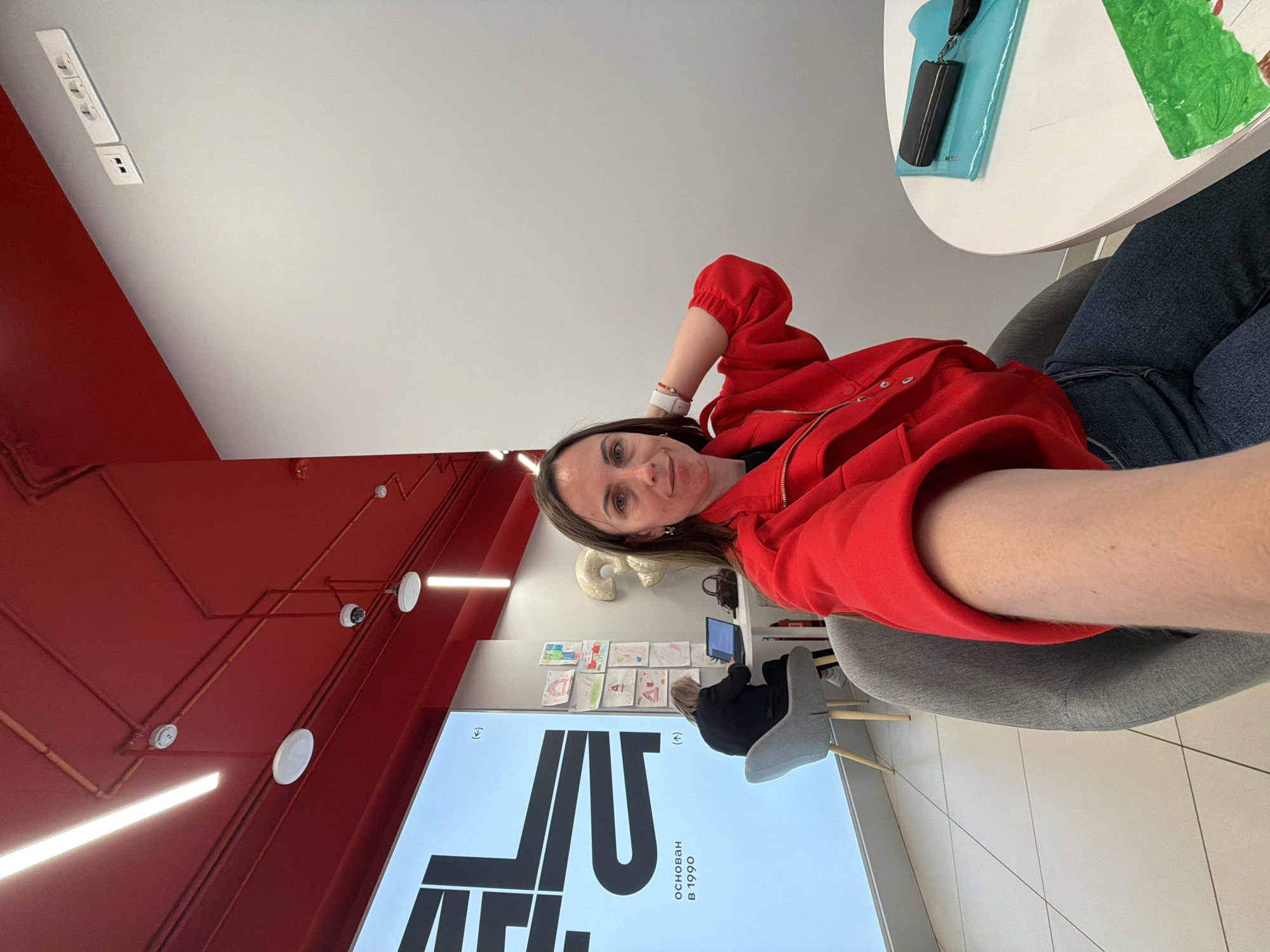 A woman in a red sweatshirt and jeans sits at a table in a bright room with red walls and ceiling structures. She is facing the camera and has her arm raised. On the table is a blue notepad and a green piece of paper. In the background, a child is seated at a computer in front of a large sign with Russian writing that says "Основан в 1990", which translates to "Founded in 1990". Artwork is displayed on the wall. Based on the writing on the sign and information available online, it is possible that this location is in Birobidzhan, Russia.