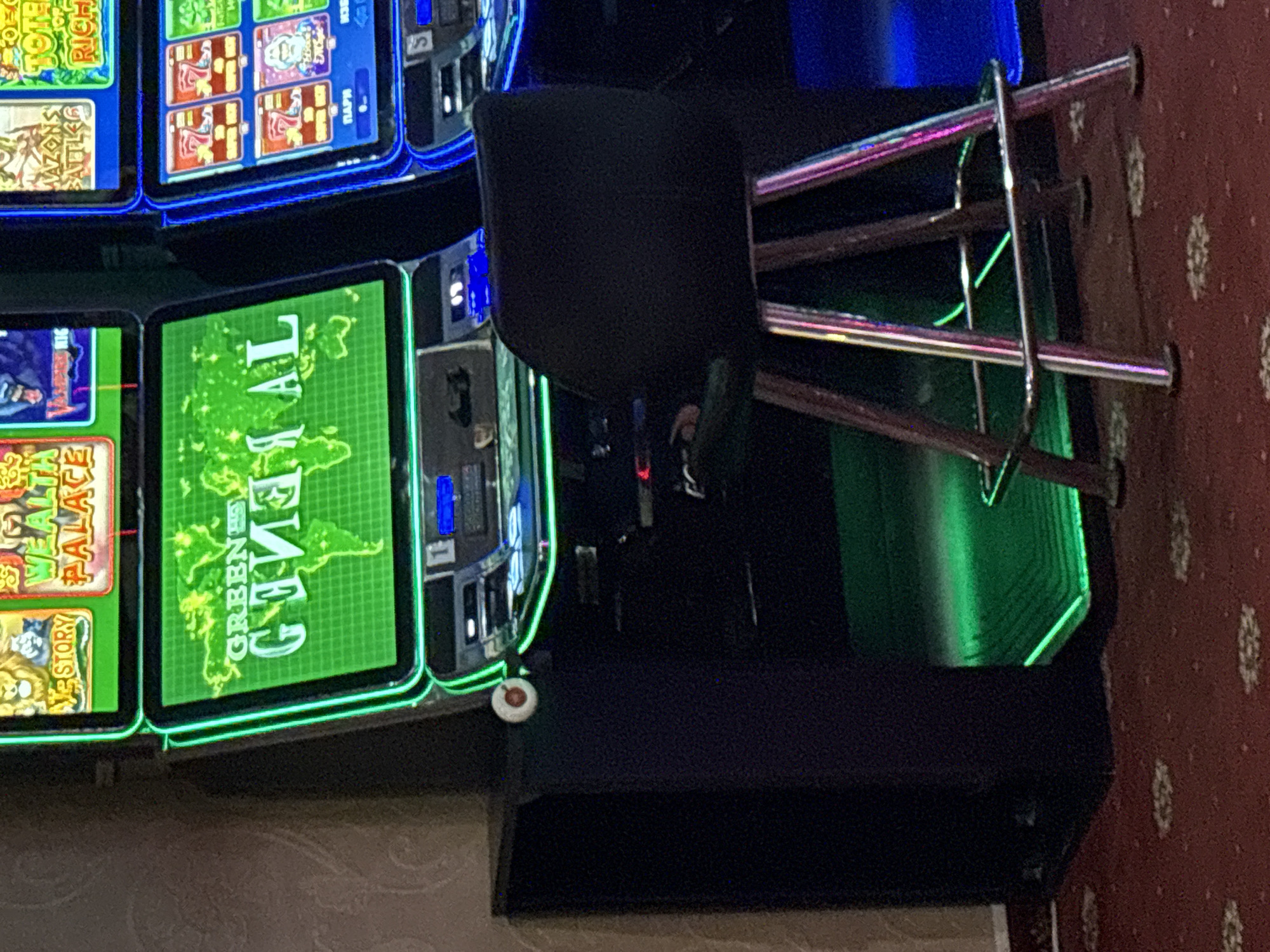 The image shows a close-up view of two slot machines in a casino setting, with a bar stool positioned in front of one of them. The slot machines feature bright, colorful displays with various game titles and graphics. One machine prominently displays a world map graphic on a green background with text that appears to read "GREEN GENIUS AL". The other machine has multiple smaller screens showing game symbols and text. The machines are illuminated with neon lights, predominantly green and blue. A dark-colored bar stool with a padded seat and metal legs is centrally located in the foreground. The floor is covered with a patterned red carpet. The background shows a textured wallpaper. The overall environment suggests an indoor entertainment venue. There are no people visible in the image. The location in the city of Karlovo, Bulgaria, is not explicitly depicted or implied by the visual content of the image.