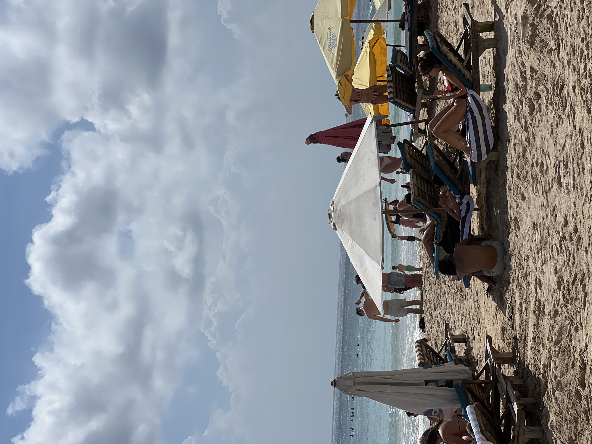 The image depicts a beach scene in Kuta Selatan, Indonesia. The sky is a mix of blue and white, with fluffy clouds. The ocean is calm, and beachgoers are visible along the shoreline. The sandy beach is populated with beach chairs and umbrellas, providing shade for sunbathers. People are relaxing, walking, and swimming in the ocean. The scene appears to capture a typical day at the beach in this location.