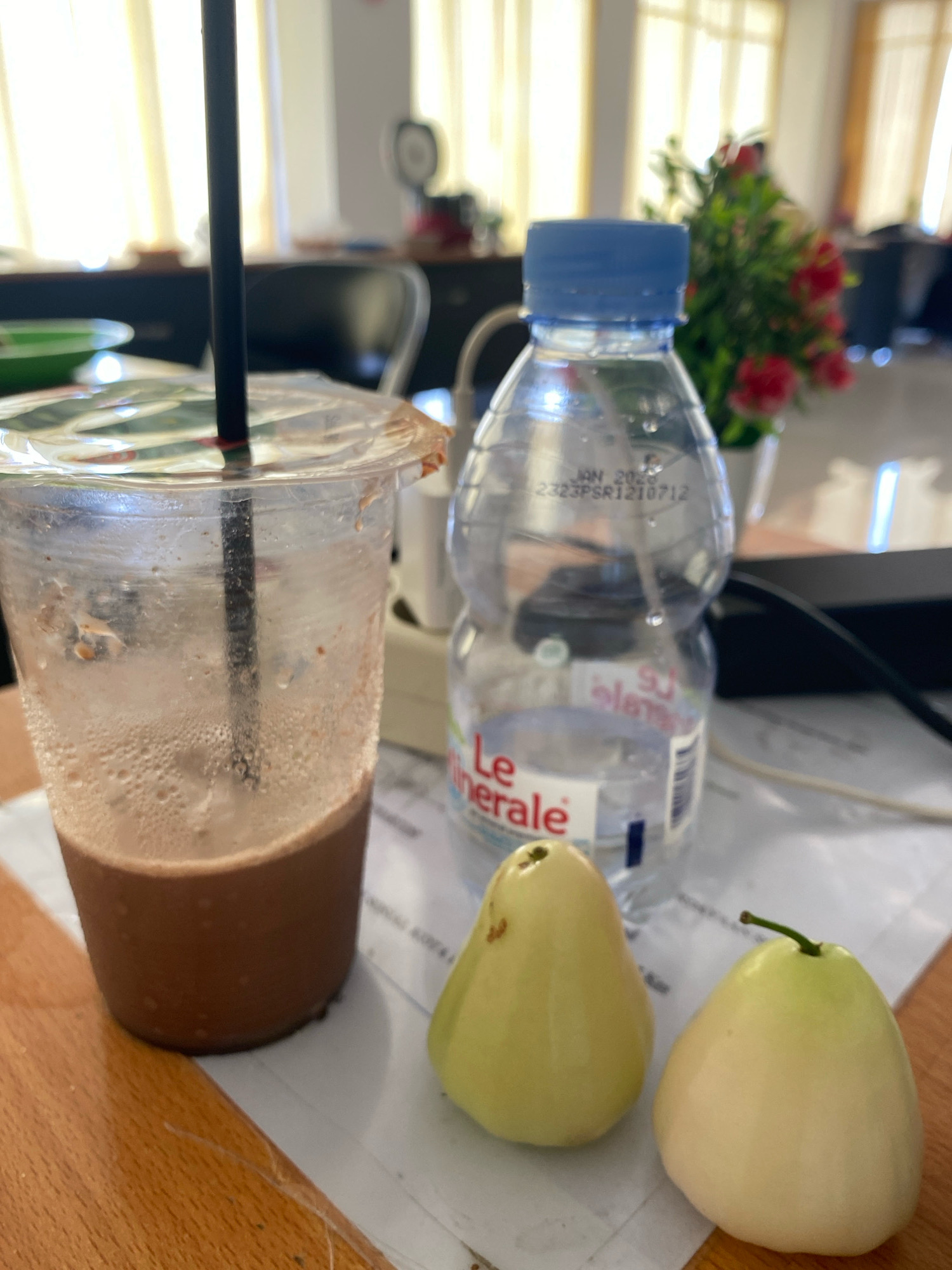 A close-up, eye-level shot captures a wooden table surface in what appears to be an office or study environment in Balaroa, Indonesia. On the table are several items: a partially consumed chocolate-flavored beverage in a clear plastic cup with a straw, a clear plastic bottle of "Le Minerale" bottled water with a blue cap, and two light green, bell-shaped fruits, possibly rose apples. The background is blurred, showing light filtering through yellow curtains, suggesting a window, and hints of office furniture and a potted plant with red flowers. The lighting is bright and diffuse, creating a soft, indoor ambiance.