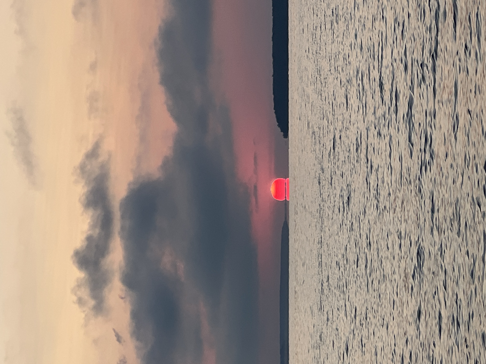 The image depicts a sunset over a body of water, with a thin strip of land visible in the distance. The sky is a gradient of soft orange and pink hues, with scattered dark clouds. The sun, a vibrant red orb, is partially submerged below the horizon, casting a warm glow on the water. The water is calm, with gentle ripples reflecting the colors of the sky. The landmass in the distance appears to be a forest or a collection of trees. The scene is serene and natural, with no signs of human activity or urban elements. The location is identified as Novopichugovo, Russia.