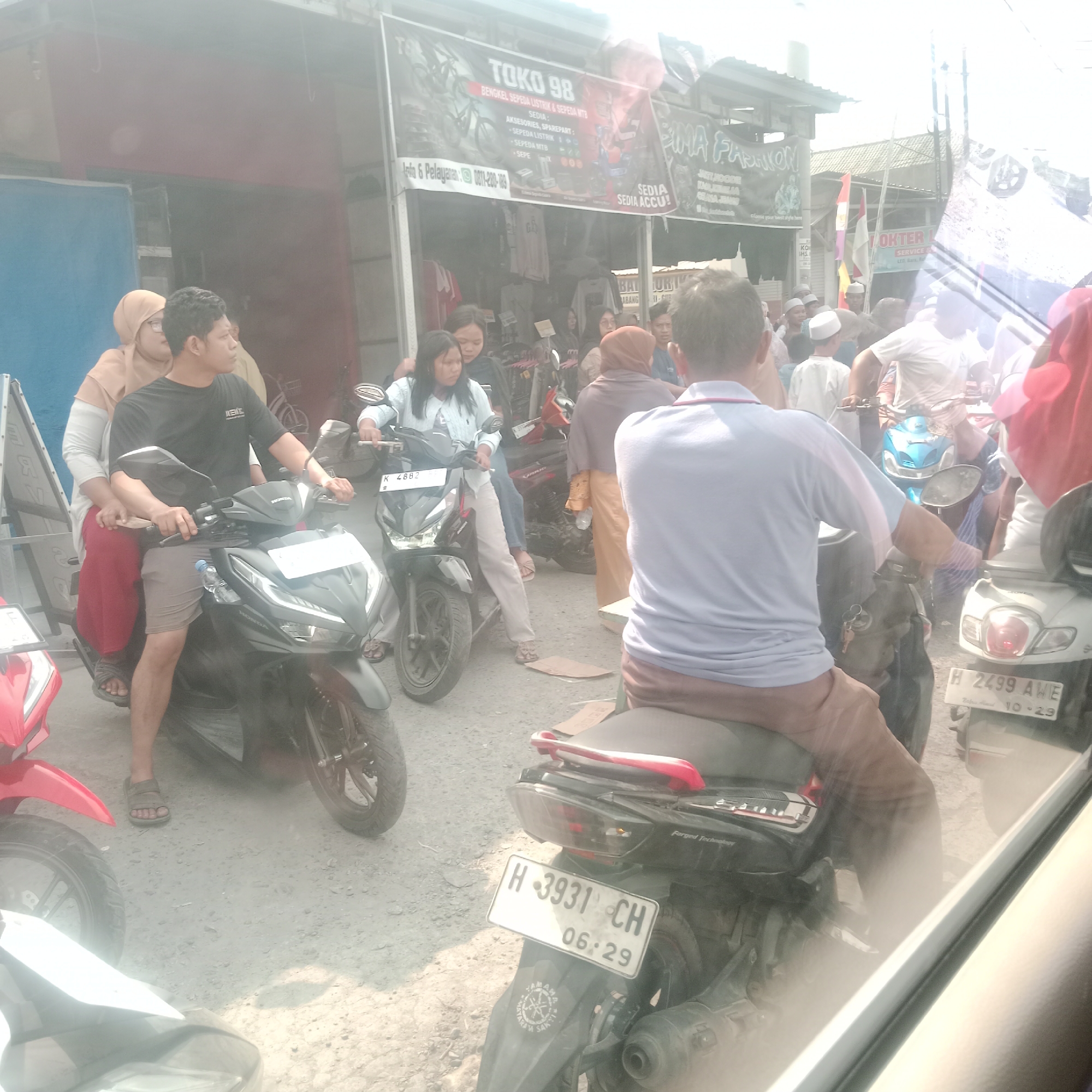 This is an eye-level, medium shot of a street scene in Jekerto, Indonesia, possibly in a market or commercial area. The foreground is dominated by several motorcycles and their riders, with a bright, hazy sky overhead.
In the left foreground, a red motorcycle's front is visible. Behind it, a dark gray motorcycle is ridden by a man wearing a black t-shirt and khaki shorts, with a woman in a peach-colored hijab and a red skirt seated behind him. This motorcycle has a license plate that reads "H-3931 CH".
To the right of this, a woman in a light blue shirt and light-colored pants is seated on a black motorcycle with the license plate "K 2882". She appears to be looking down. Next to her, other individuals are visible, some on motorcycles, including one with a blue scooter.
In the background, there are several storefronts with banners. One banner, above a shop, reads "TOKO 98", with text below indicating it sells bicycle spare parts and accessories. Another banner displays "SEMA FASHION". Further to the right, a sign for "OKTER" is visible, possibly indicating a service or business. The ground is unpaved and dusty, typical of a street in a developing area. The overall lighting suggests it is daytime with strong sunlight causing a bright, washed-out effect in some areas. The presence of multiple motorcycles suggests traffic or a gathering of people on their vehicles.