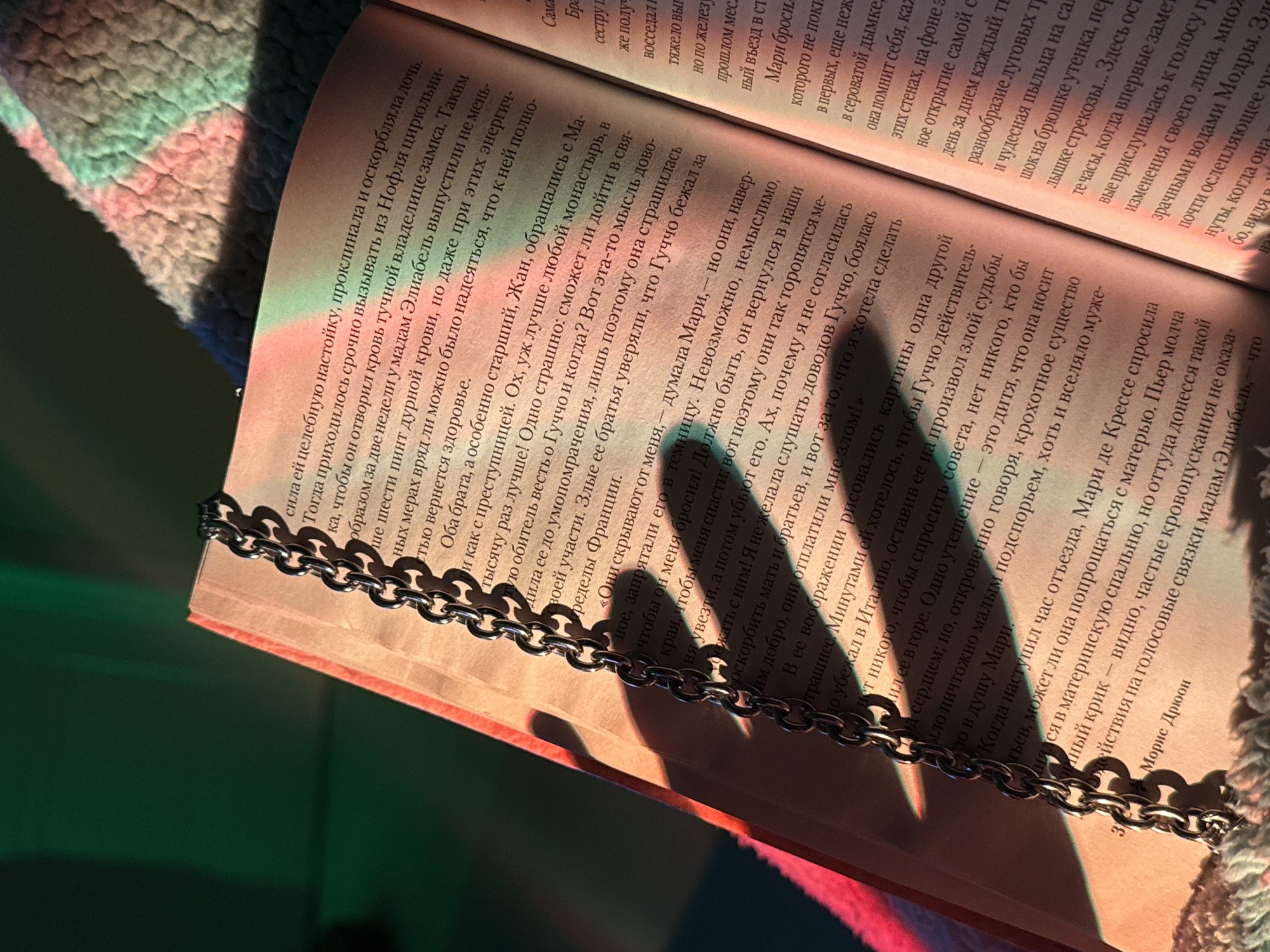 Here is a forensic description of the image:
The image is a close-up shot of a book, open and lying on a surface that is out of focus. The pages of the book are filled with text in what appears to be the Russian language. The book is bound in a reddish cover. Draping over the open pages of the book is a metal chain, casting shadows on the text beneath.