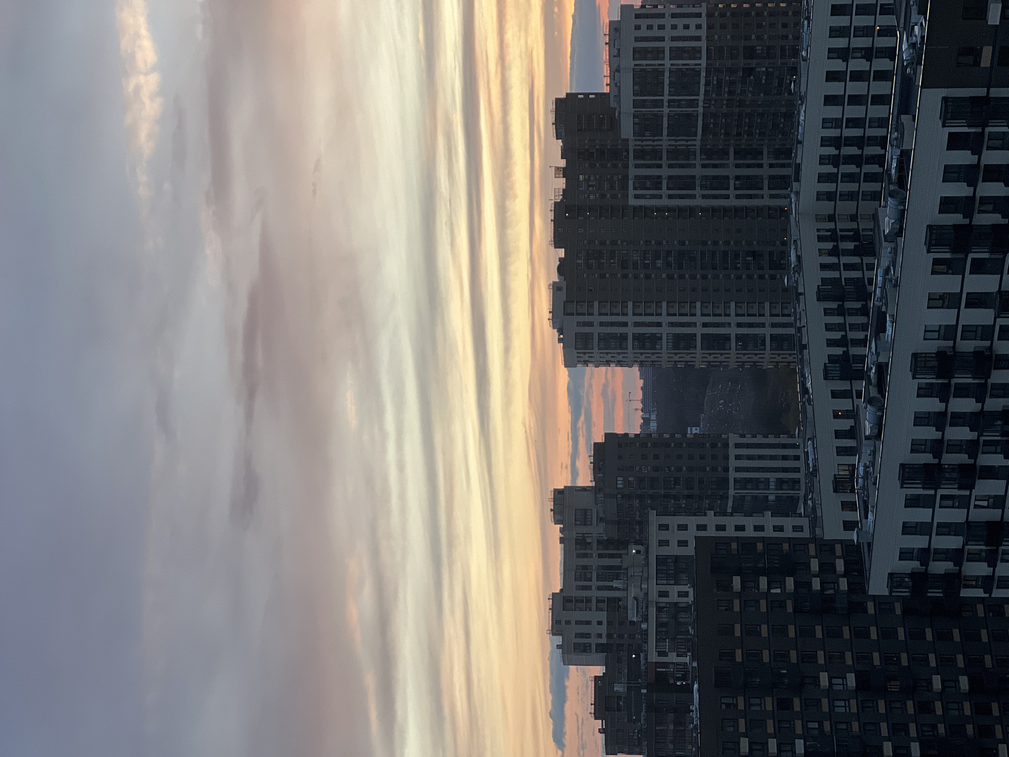 The image depicts a cityscape at dusk, likely in Khimki, Russia, judging by the architectural style of the buildings. A line of densely packed, modern apartment buildings with a repetitive pattern of windows and balconies are the dominant structures. The buildings are varying shades of gray and black, suggesting different materials or stages of construction. The sky exhibits a striated cloud formation, with hues of gray, orange, and yellow indicating the setting sun. A highway or road is faintly visible between some of the buildings in the middle ground. There are no visible people or explicit actions in the frame. The image is rotated 90 degrees clockwise.