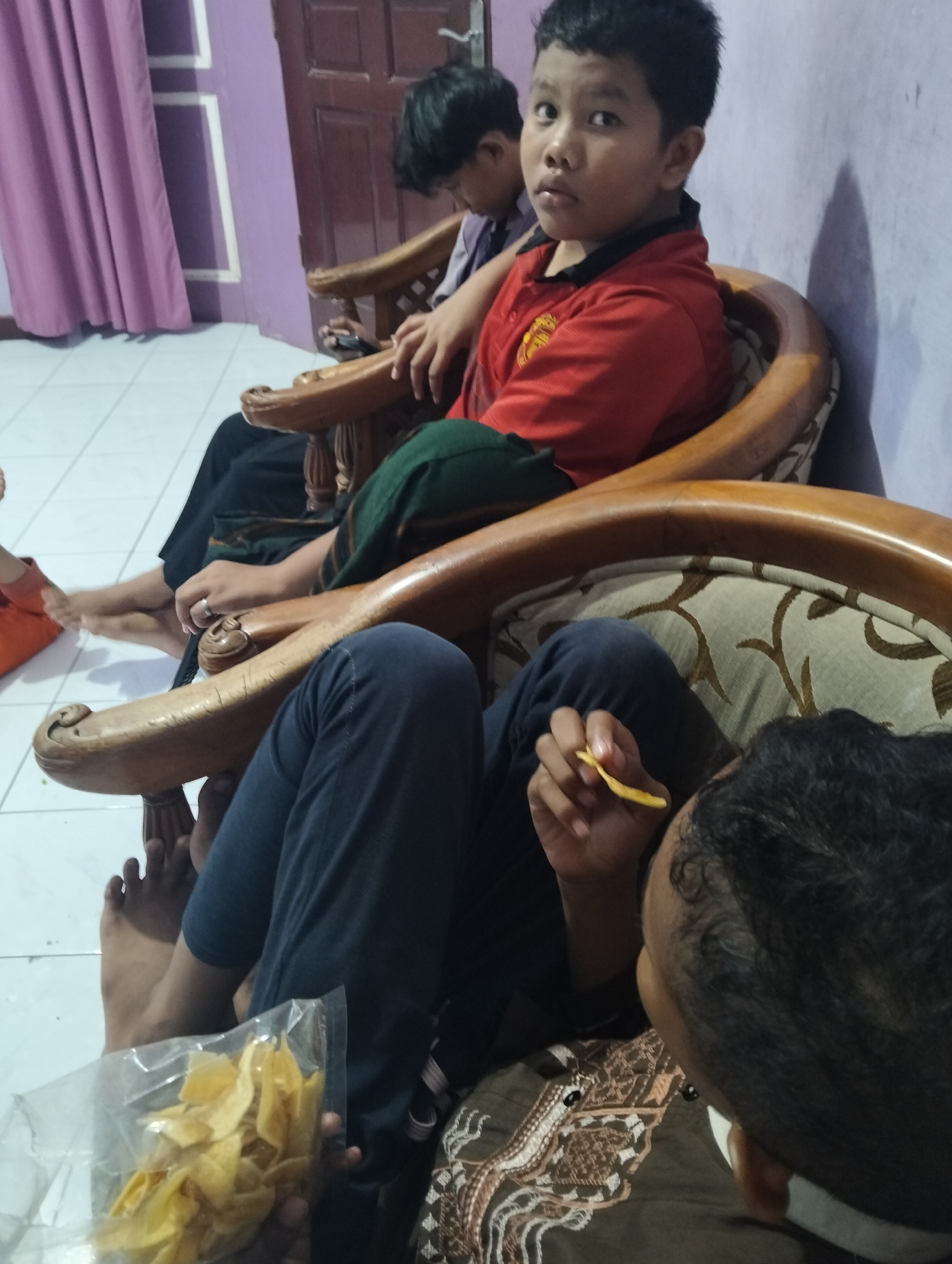 The image depicts a group of children and adults in what appears to be a residential setting in the city of Medan, Indonesia. In the foreground, a young boy is seated on a wooden armchair, wearing a red shirt with a black collar. He is looking towards the camera with a serious expression. Next to him, a person is partially visible, holding a bag of what seems to be banana chips and a single chip in their hand, suggesting they are eating. Further back, other individuals are seated, with one child in a purple shirt visible in the background. The environment includes a purple curtain on the left, and a brown wooden door on the right, possibly within a living room or dining area. The floor is tiled. The overall scene suggests a casual gathering.