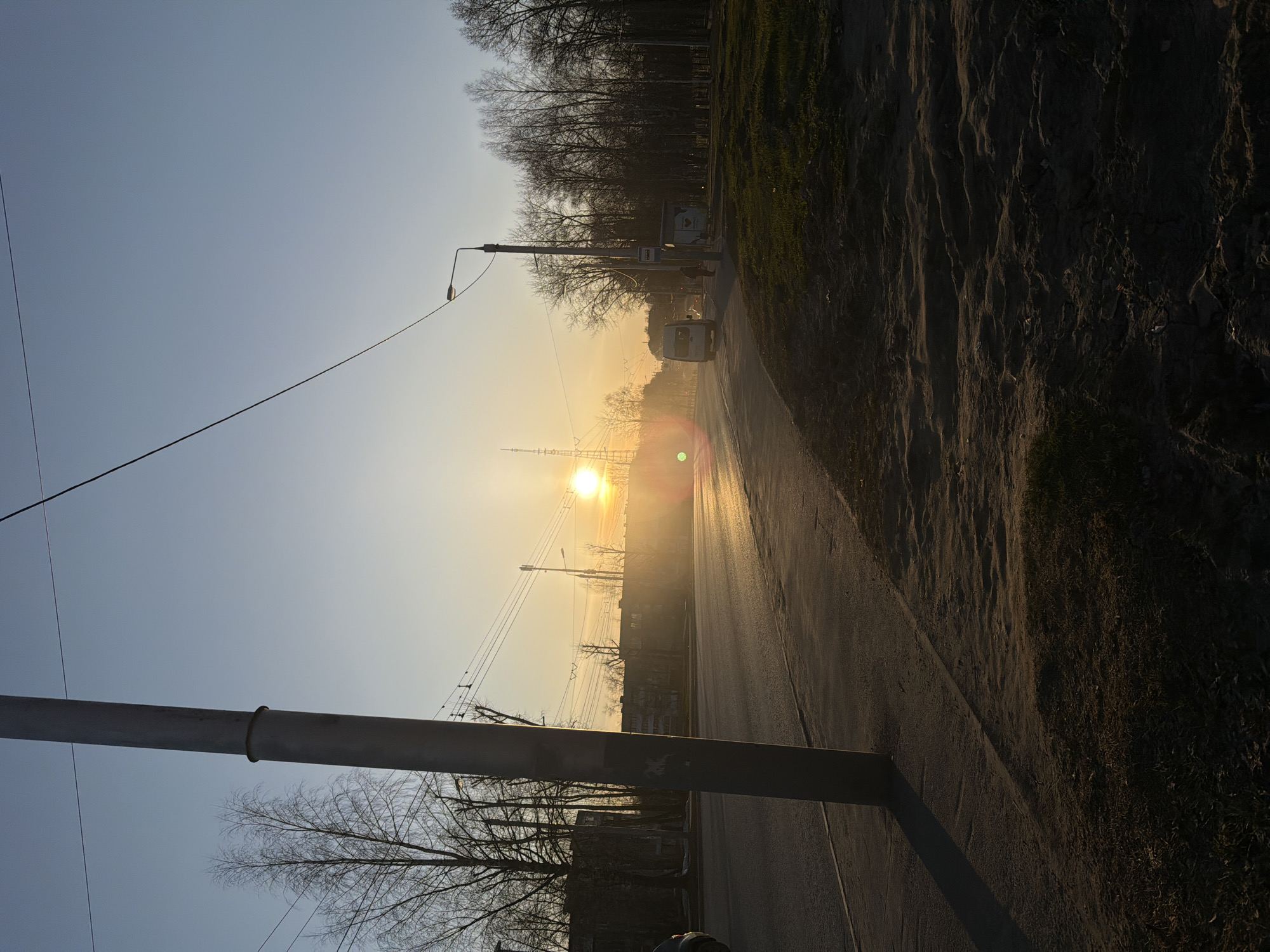The image shows a sunlit street in Berezniki, Russia. The scene is dominated by the road receding into the distance, with the sun positioned directly ahead, causing a strong glare. The road appears to be asphalt. On the right side of the road is a dark, sloped area, likely a roadside embankment or a patch of vegetation. On the left, there is a utility pole with wires extending across the sky. Bare trees are visible on both sides of the road, indicating a winter or early spring season. A vehicle, potentially a van or a small truck, is visible further down the road. A bus stop is visible near the vehicle. The sky is a pale blue color, brighter near the sun. The overall impression is that of a late afternoon or early morning scene due to the low angle of the sun.