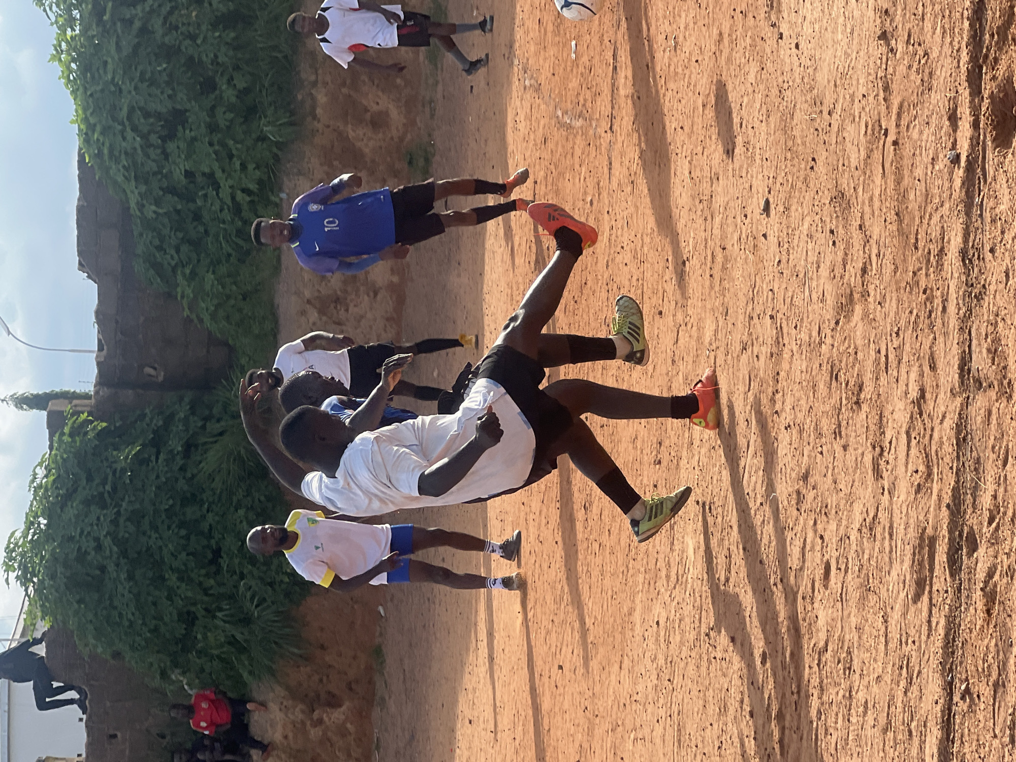 In Jos, Nigeria, a group of young men are engaged in a casual game of soccer on a dusty, reddish-brown dirt field. The sun casts long shadows across the uneven terrain. In the foreground, two players are in the midst of action. One player, wearing a white t-shirt and black shorts, is in motion, his right leg extended as if to kick a ball, which is partially visible to the right of the frame. His left leg is bent, supporting his weight. The other player, also in a white t-shirt and black shorts, is positioned behind and to the side, his body angled as he appears to be reacting to the play.
In the background, several other individuals are present. To the left, a man in a white t-shirt and blue shorts stands observing. Further back, a player in a blue jersey with the number 10 on it is visible, wearing black shorts and black socks. Another player in a white t-shirt and black shorts is seen in the far right background. To the left of the field, a man in a red shirt is seated on a raised embankment. On the top of the embankment, a person in a black hooded jacket is seated. The environment is characterized by sparse vegetation and some architectural structures in the distance, suggesting an urban or semi-urban setting within Jos.