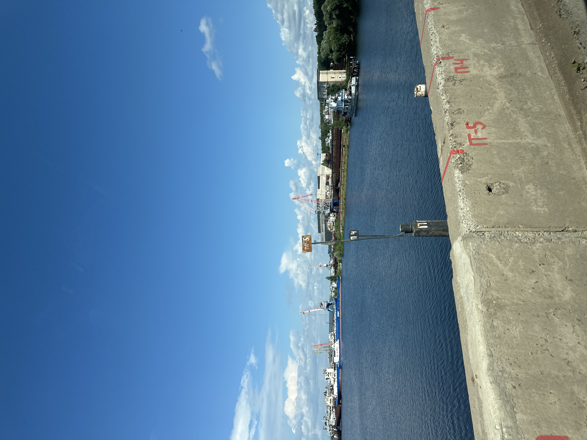 The image depicts a wide river with a docked fleet of ships and industrial cranes on the far bank. The sky is a vibrant blue with scattered white clouds. In the foreground, a concrete barrier is visible, marked with red inscriptions. On the water, a navigational marker with red and white markings is present. The scene appears to be a port or shipyard, likely located in the city of Chaikovsky, Russia, given the context of the prompt. The overall atmosphere is industrial and utilitarian under a clear summer sky.