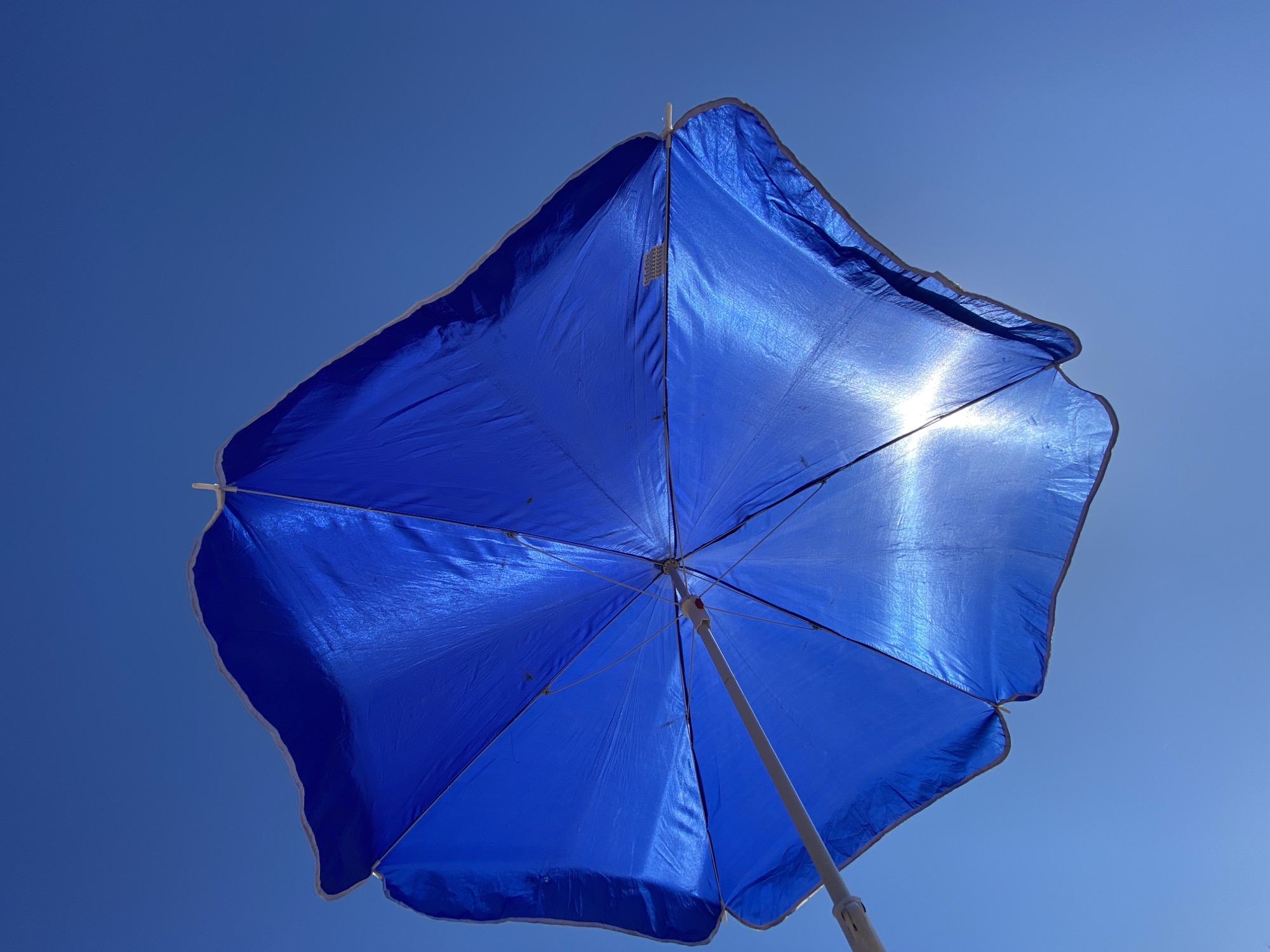 The image displays a royal blue beach umbrella, viewed from directly underneath, against a clear, azure sky. The umbrella fabric is slightly translucent, revealing its structural ribs and showing distinct folds and wrinkles. Sunlight, originating from behind and to the lower right, creates a bright lens flare that illuminates a section of the umbrella, highlighting its texture and giving the fabric a luminous quality. A light-colored central pole extends downwards from the umbrella's canopy. A small, rectangular label or tag with indistinct text is affixed to one of the upper panels of the umbrella. The edges of the umbrella panels feature a thin, light-colored trim. No individuals or additional objects are present in the frame. The scene indicates an outdoor environment under bright, clear weather conditions in the city of Unknown, Unknown.