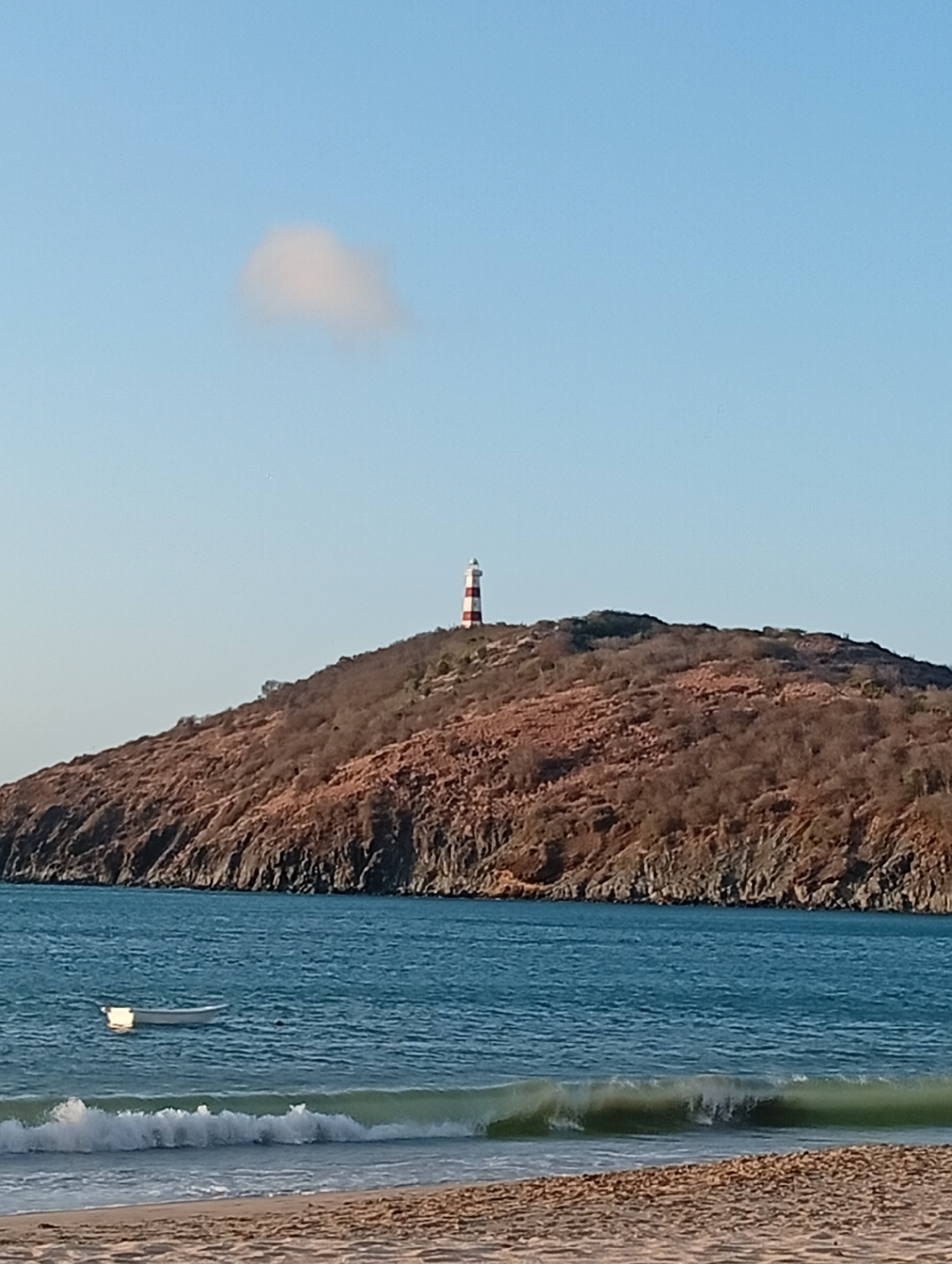 The image depicts a coastal scene near Pedro González, Venezuela. A red and white striped lighthouse sits atop a brown, hilly island. The island has sparse vegetation, with many bare trees. The water is blue-green, with small waves breaking on a sandy beach in the foreground. A small, white boat is anchored in the water, closer to the island. The sky is light blue with a single small, white cloud visible above the lighthouse. The scene appears to be taken during the daytime with good visibility.