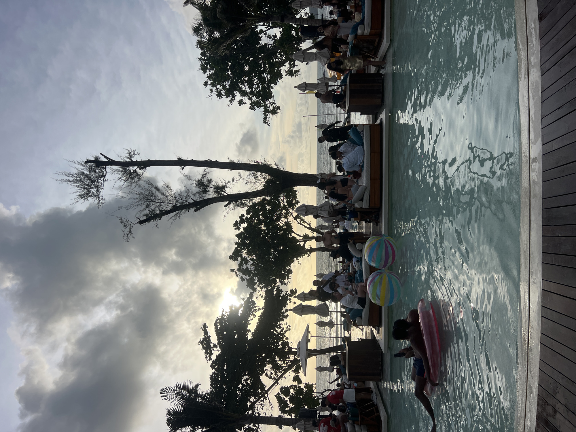 The image depicts a crowded beach club in Patong, Thailand, during a cloudy sunset. In the foreground, a person lies on a pink inflatable raft in a swimming pool, holding a phone. Two large, striped beach balls float nearby. The midground shows several people lounging on daybeds, some dressed casually, others in swimwear. Behind them, the beach is visible with umbrellas, more daybeds, and a crowd of people. The ocean stretches to the horizon, with the sun partially obscured by clouds, casting a warm glow. Palm trees and other tropical foliage frame the scene. The overall atmosphere appears to be relaxed and social, with a mix of people enjoying the end of the day at the resort.