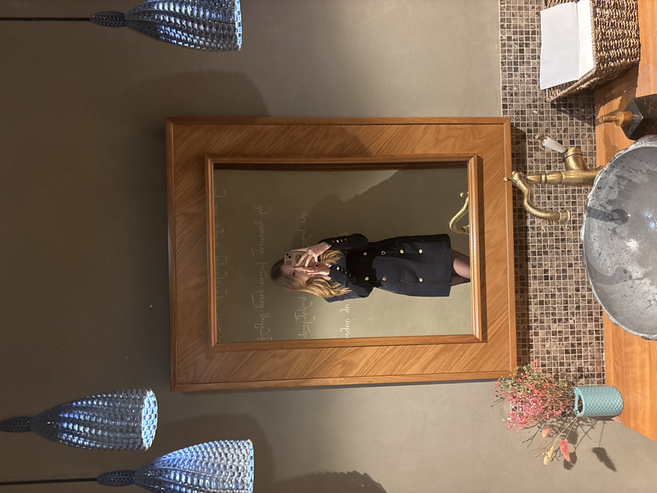 This image depicts a female subject taking a mirror selfie in a restroom or washroom facility. The location is identified as Hinkalnaya No.1 Restaurant, situated at Ulitsa Voroshilova, 137, Serpukhov, Russia.
The primary subject, reflected in a large rectangular mirror with a thick light-wood frame, is a woman with long, light-brown hair. She is wearing a dark, double-breasted dress featuring two vertical rows of gold-colored buttons, along with dark tights. Her hands are raised, holding a light-colored smartphone to capture the reflection.
Above the mirror, two light blue, woven-textured pendant lamps hang from the ceiling, illuminated from within. The walls are a uniform, plain olive-green or dark beige color. Directly behind the woman's reflection, on the wall inside the mirror's view, several lines of cursive Georgian script are visible, including the phrase "გემრიელი ბროწეულის წვენები" (Delicious pomegranate juices).
Below the mirror, a wooden countertop supports a dark, irregular-shaped stone basin. To the right of the basin, an antique-style brass faucet with a white handle accent is installed. Further to the right on the countertop, a dark, rectangular soap dispenser with a brass pump is positioned next to a woven brown basket containing folded white paper towels. To the left of the basin, a small teal vase holds a collection of dried flowers and twigs, predominantly in shades of pink and muted brown. The wall immediately beneath the countertop is covered in small, square mosaic tiles in various shades of brown and beige.