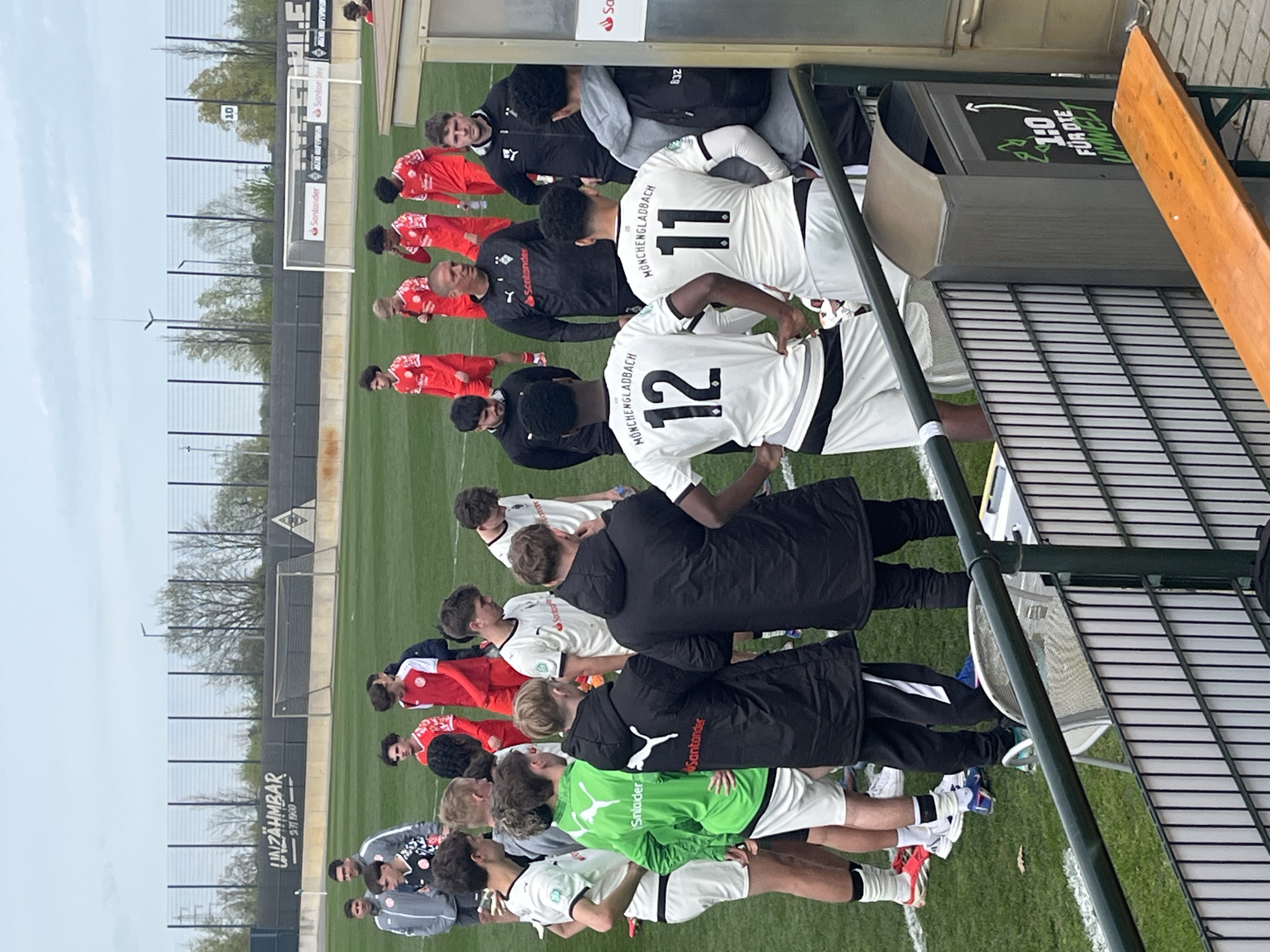 The image depicts a soccer field in Mönchengladbach, Germany. A soccer team wearing white and black uniforms with the word "Mönchengladbach" printed on the back stands alongside the field's edge. They are next to a bench and other objects. Another soccer team wearing red and white uniforms stands adjacent to the other team. The environment is an outdoor soccer field with advertising along the perimeter.