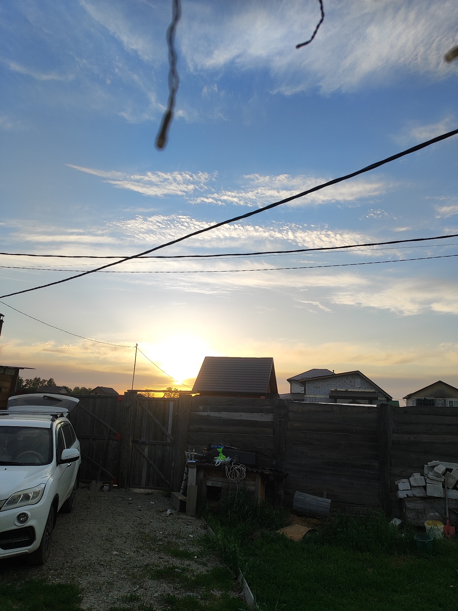The image captures a residential or semi-rural scene at sunset in Marusino, Russia. The sky, prominent in the upper two-thirds of the frame, displays a gradient of colors from clear blue at the top, transitioning to lighter blues, whites, and hues of orange and yellow near the horizon. Scattered white clouds, some wispy and others more textured, are distributed across the sky, with the most vibrant coloration occurring near the sun.
The sun is visible as an intense, bright light source on the horizon, slightly to the right of the central vertical axis, partially obscured by the roofline of a house. Several dark utility lines, appearing as horizontal and diagonal strands, traverse the sky in the mid-ground. Two distinct, dark, blurred, wavy objects, likely close to the camera lens (e.g., hair or a thin wire), are visible in the upper portion of the frame, one vertically oriented near the center-right and a smaller one near the top-right corner.
The lower third of the image shows ground features and structures. On the left, a white SUV, possibly a crossover model, is parked on a gravel surface. The vehicle's trunk lid appears to be open, and a roof rack is mounted. The ground in this area is primarily gravel with patches of green grass. A tall, dark wooden fence, constructed from horizontal logs or planks, extends across the mid-ground, separating the foreground from the background. A wooden gate, matching the fence material, is situated to the right of the SUV.
Behind the fence and gate, a series of houses are visible. One prominent house, silhouetted against the sun, features a dark, sloped roof. To its right, another house is visible, constructed with light grey concrete blocks and showing exposed framing or unfinished walls, indicating ongoing construction. Further to the right and left, additional residential structures with various roof types are partially visible. Sparse tree foliage extends above the fence line, particularly to the left of the sun's position. Adjacent to the wooden gate on the right, a makeshift workbench or stand holds miscellaneous items, including coiled cables and green plastic objects. Further right along the fence, a stack of grey concrete blocks, a shovel, and two plastic buckets (one green, one yellow) are present on the ground. No individuals are visible in the scene.