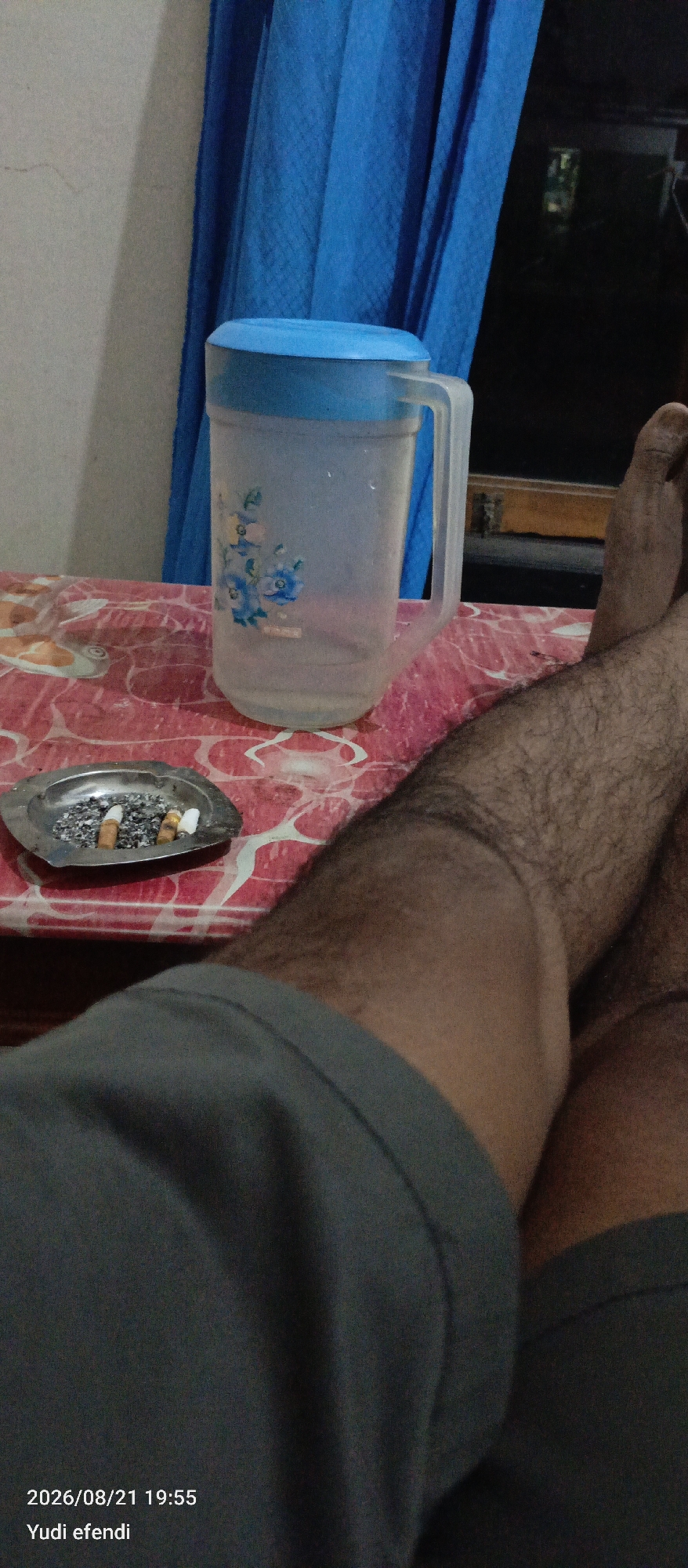 The image depicts an indoor scene with a focus on a table covered with a red and white patterned cloth. On the table, there is a transparent pitcher with a blue lid and floral decoration, a metal ashtray containing several cigarette butts, and a person's legs and feet, covered in dark hair. The person appears to be sitting or lying down, with their legs extending into the foreground. To the left of the table, a blue curtain is visible against a light-colored wall. The background is dark, suggesting it might be nighttime or an area with limited lighting, with some indistinct shapes visible through a window. A timestamp and name, "2026/08/21 19:55 Yudi efendi," are overlaid on the bottom left of the image. The location of Rawajitu Selatan, Indonesia, is provided as context.