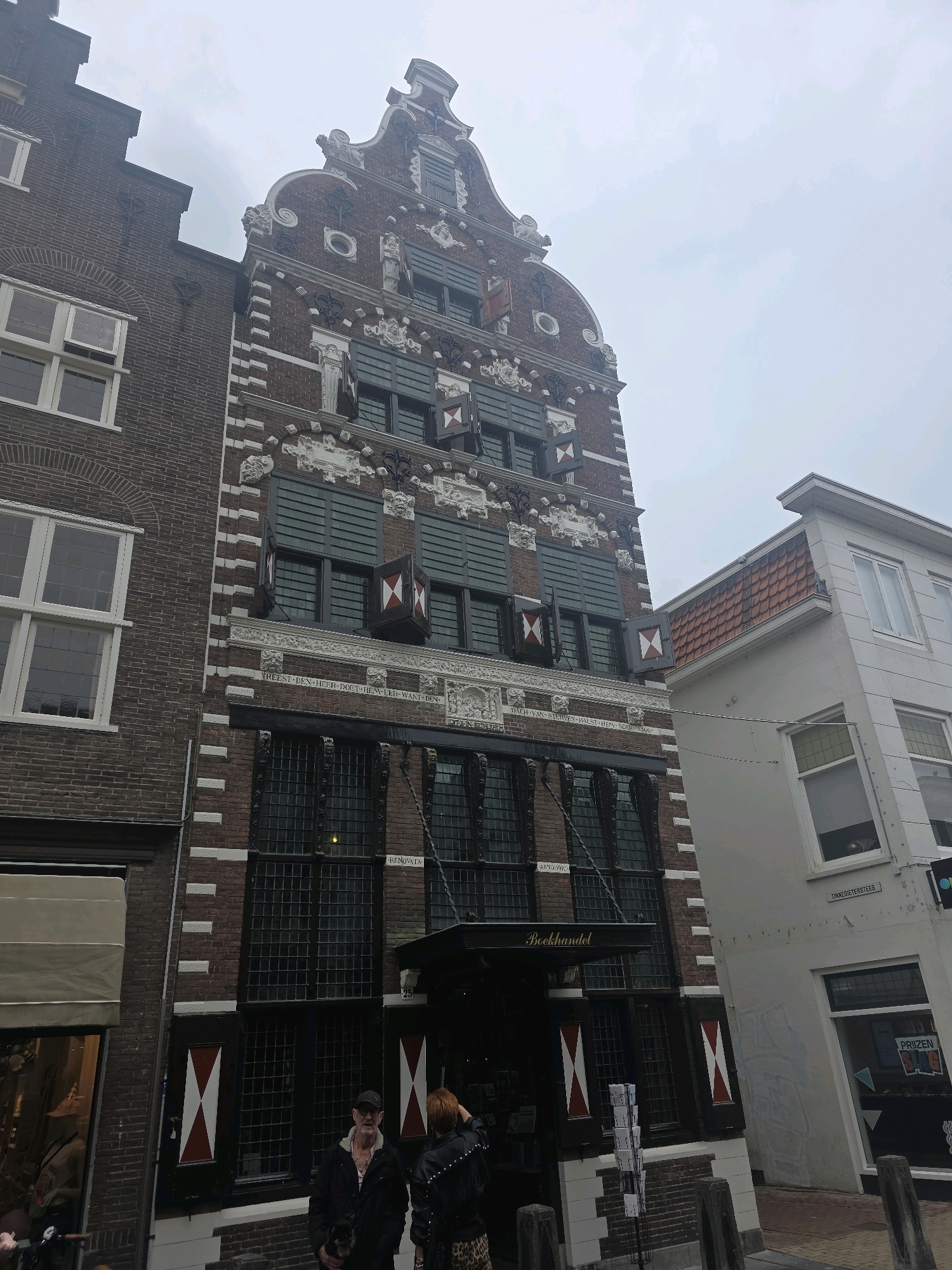 The image shows a street scene in Gorinchem, Netherlands, featuring the facade of a historic building with ornate architecture. The building is primarily brick with white decorative elements and large, multi-paned windows. It has a stepped gable roofline typical of Dutch architecture. On the facade, there are several inscriptions and decorative carvings. The ground floor appears to be a shop with the name "Boekhandel" (bookstore) visible above the entrance. Two individuals are standing in front of the bookstore, one man and one woman. The man is wearing a dark jacket and a baseball cap, holding a small dog. The woman is wearing a black leather jacket with studs and a leopard-print skirt. The street is paved, and there are a few street elements like a bike rack and planters visible. The sky is overcast.