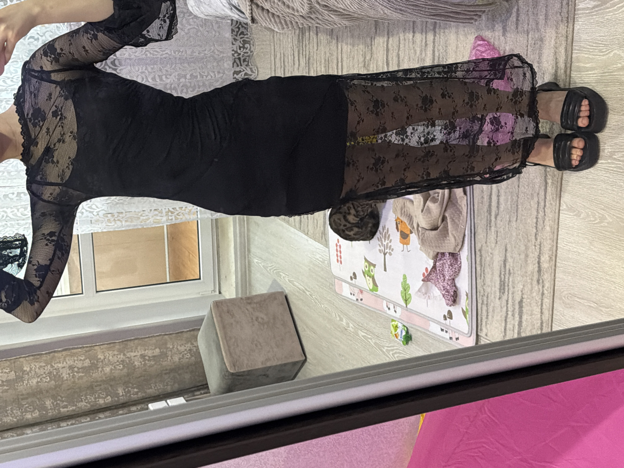 A photograph shows a person wearing a black lace dress and platform sandals. The person is indoors, near a window, with a grey stool nearby. A play mat with cartoon designs is on the floor. A piece of furniture is partially visible in the background.