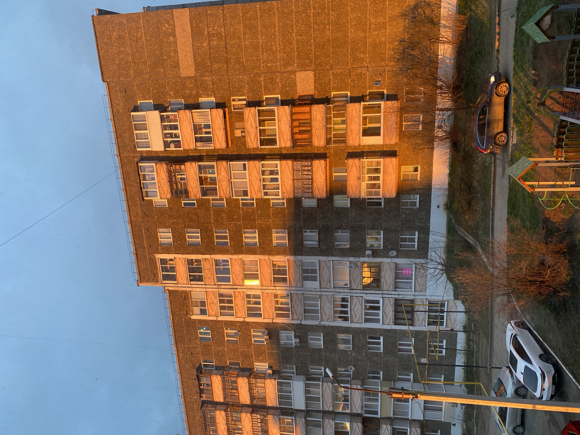 The image captures a residential street scene in Votkinsk, Russia, bathed in the warm, golden light of either sunrise or sunset. The primary subject is a large, multi-story concrete apartment building, characteristic of Soviet-era architecture, stretching across the frame. Its facade is textured and features numerous windows and enclosed balconies, many of which have wooden shutters or blinds. The sun illuminates the upper and middle sections of the building in vivid orange and yellow tones, contrasting with the cooler grey of the lower, shadowed parts.
On the ground level, a paved pathway runs alongside the building, where two cars are parked. A dark-colored car, possibly blue or black, is visible near a small playground equipped with green and orange structures. Further along, a white car is partially visible. The ground is covered with sparse vegetation and bare-branched trees, suggesting late autumn or early spring. Utility poles and power lines are present, with one prominent yellow pole and associated wires in the foreground. The sky is clear and a pale blue, with a single dark wire stretching across it.
Notable details include the dramatic lighting that highlights the building's texture and the reflections in many windows. Inside one apartment on a lower floor, a pink neon light, possibly a stylized letter, glows brightly. No specific human activity is evident. The overall atmosphere is tranquil, captured during a transitional period of the day. No clear, legible text is visible other than the potential neon light.