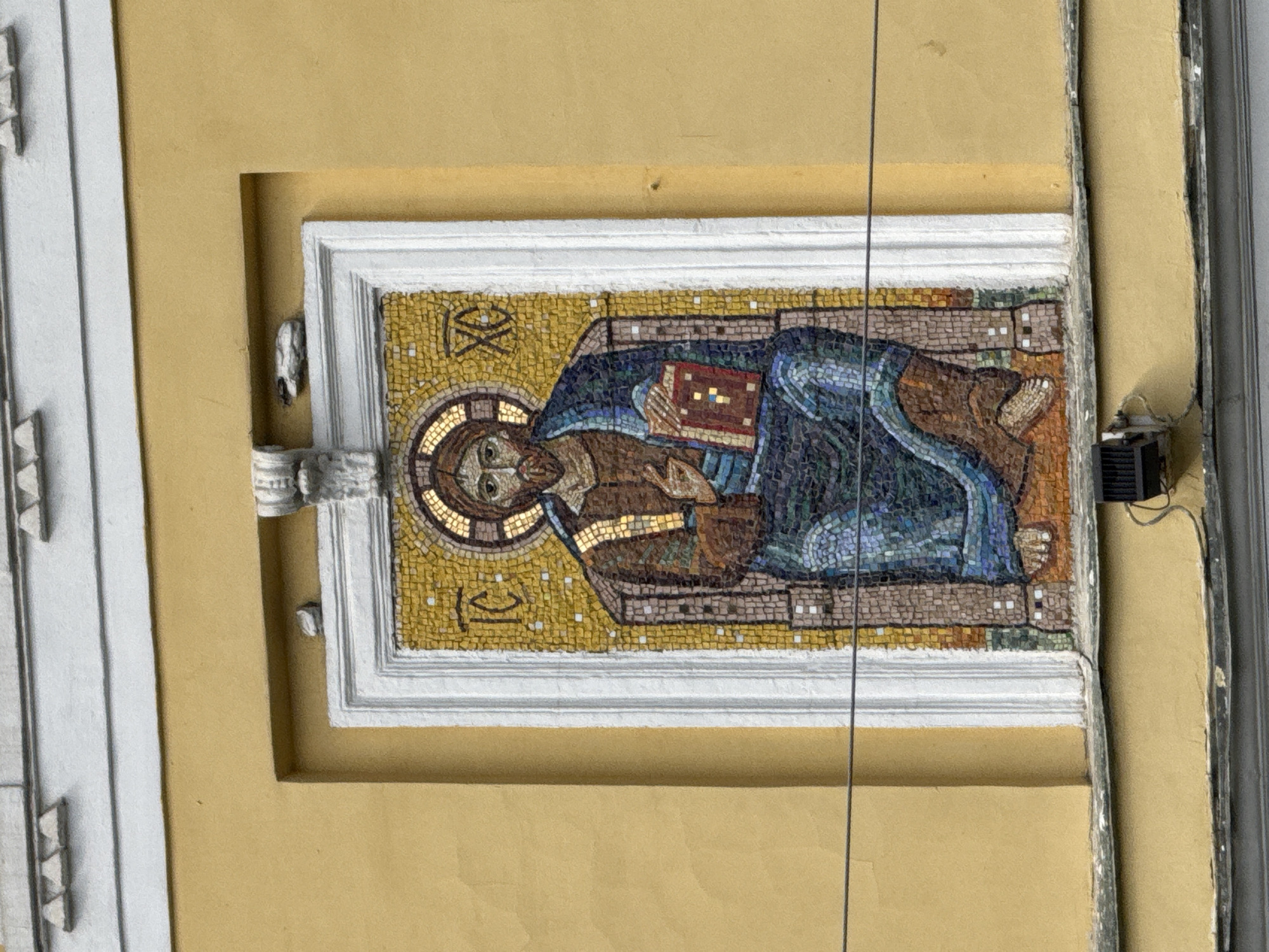 This is a medium shot, eye-level, exterior photograph of a mosaic of Jesus Christ on the side of a yellow building in Moscow, Russia. The mosaic is framed by a white, ornate border and is set within a rectangular recess. Jesus is depicted as a bearded man with a halo, seated on a throne-like chair, holding an open book. His robes are depicted in shades of blue and brown. To the left of his head are the Greek letters "IC" and to the right are the letters "XC", which are abbreviations for Jesus Christ. The building's facade is painted a light yellow, and the top of the image shows a white architectural detail with triangular projections. A thin black wire runs horizontally across the lower portion of the mosaic. There are also some small, light-colored stones affixed to the wall above the mosaic. Below the mosaic, a dark gray metal box with a vent is visible, attached to the wall with wires. The bottom of the image shows another architectural element of a darker gray color.