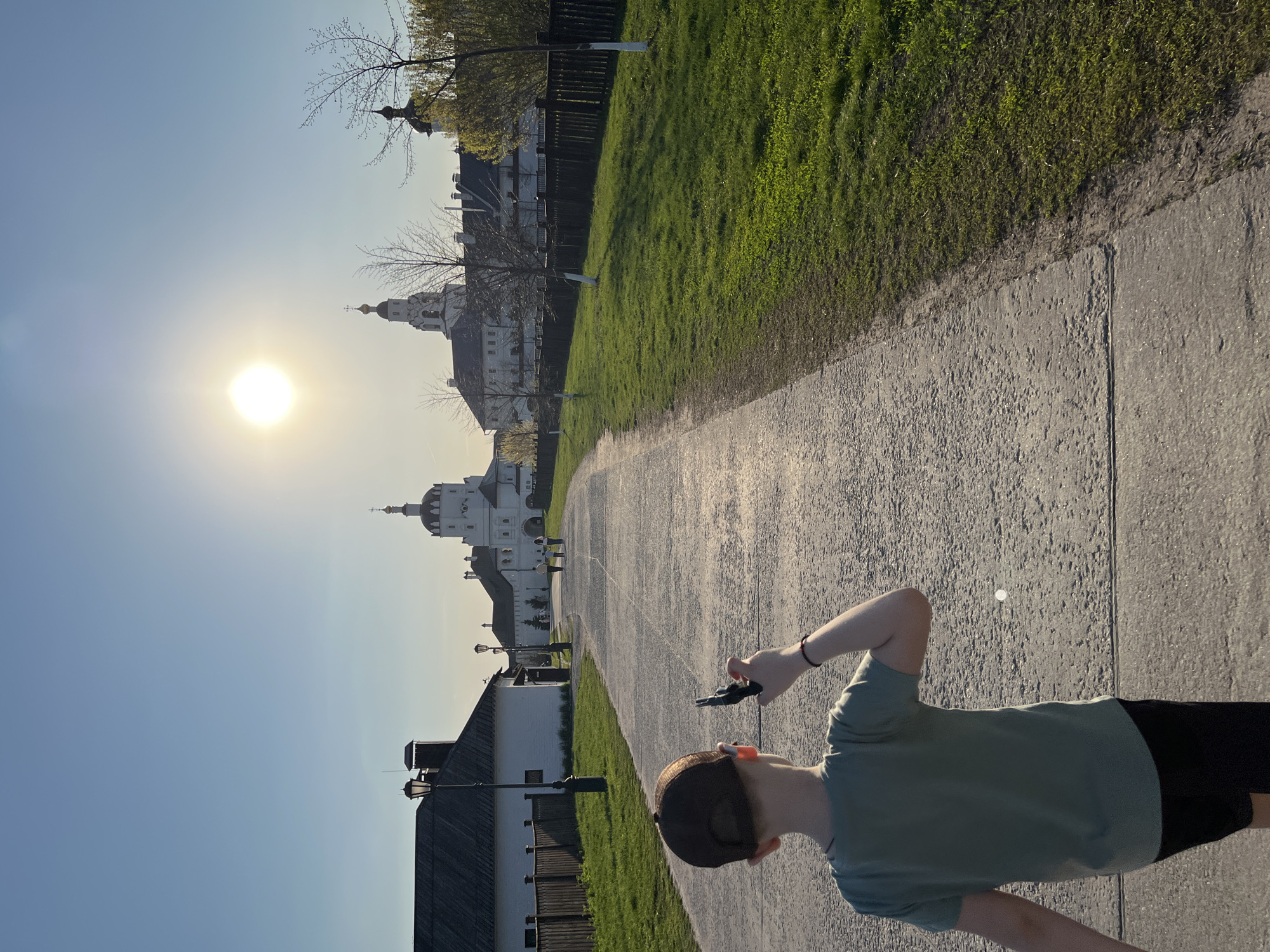 The image shows a person walking down a paved pathway in Sviyazhsk, Russia. The person, wearing a green t-shirt and dark shorts, holds a small object in their hand. The pathway leads towards a group of white buildings with domes, likely a monastery or church complex, bathed in sunlight. A green grassy area borders the path on one side, and a wooden fence and other buildings are visible on the other side. The sky is clear and blue.