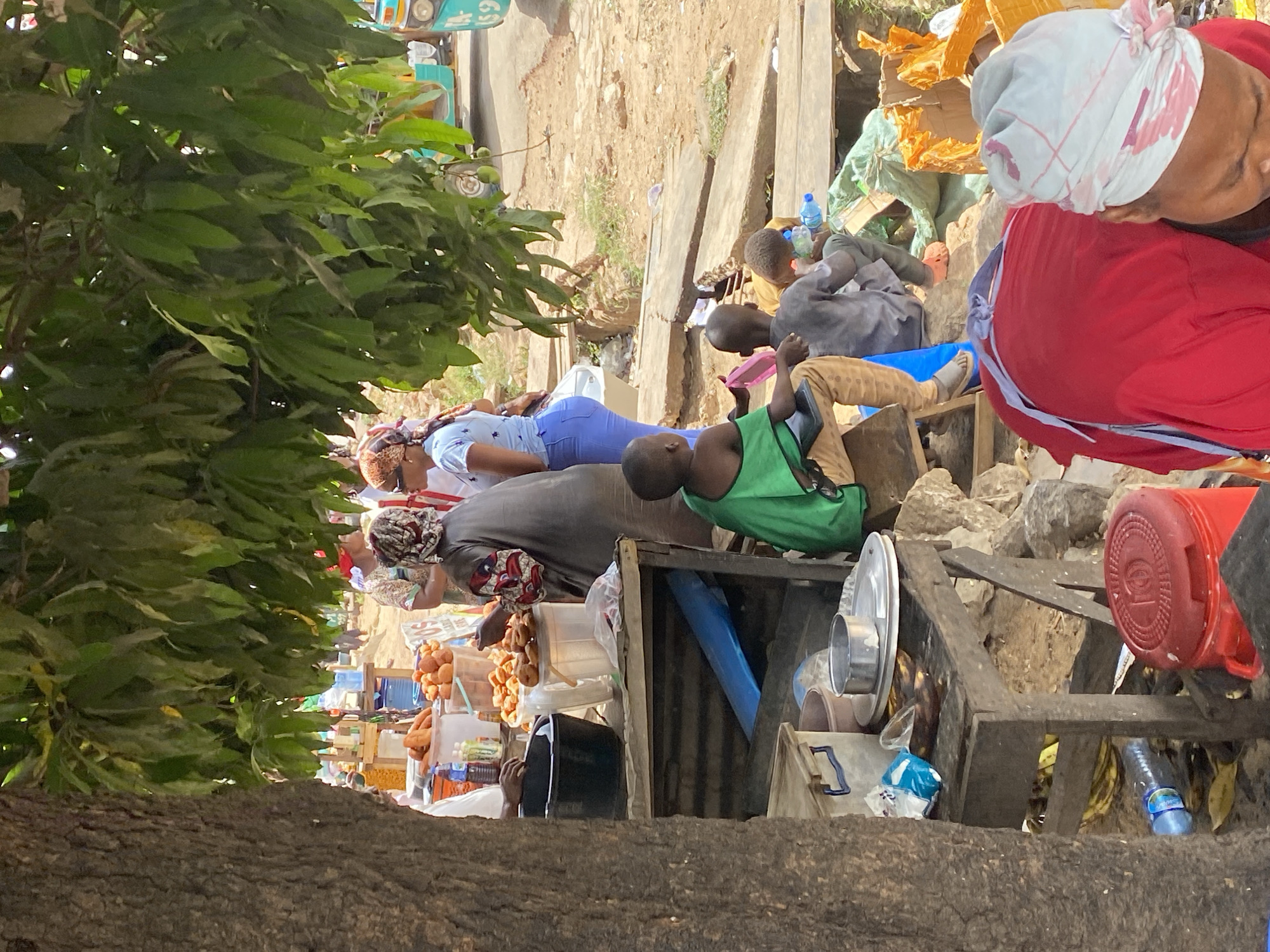 This image captures a vibrant, informal outdoor market scene, likely in Jos, Nigeria, given the provided context. The view is partially obscured on the left by the thick foliage and trunk of a large tree. The setting appears sunny and dry, with dusty, uneven ground forming the marketplace.
Several people, including adults and children, are visible amidst makeshift market stalls constructed from wood and various materials. In the foreground, a person in a light blue top and jeans is lying down, possibly resting. To their right, a woman in a dark grey top and patterned headscarf attends to a stall displaying round, golden-brown food items, possibly fried snacks like akara or puff-puff, in clear containers. A small sign with the number "50" is visible nearby.
Further to the right, a child in a green top sits on a wooden structure, looking forward, while another child in a grey outfit sits nearby. In the upper right corner, a person wearing a red top and a white and red patterned headscarf is seen from the back. Various objects are scattered around the stalls, including plastic water bottles, metal bowls, a red plastic bucket, cardboard boxes, and blue tarps or containers.
In the background, beyond the immediate market activity, more dusty ground is visible, and a light blue commercial vehicle, possibly a bus or truck, with the numbers "69" visible on its side, is parked. The overall scene depicts a bustling, everyday market environment, characterized by informal commerce and community interaction under bright sunlight.