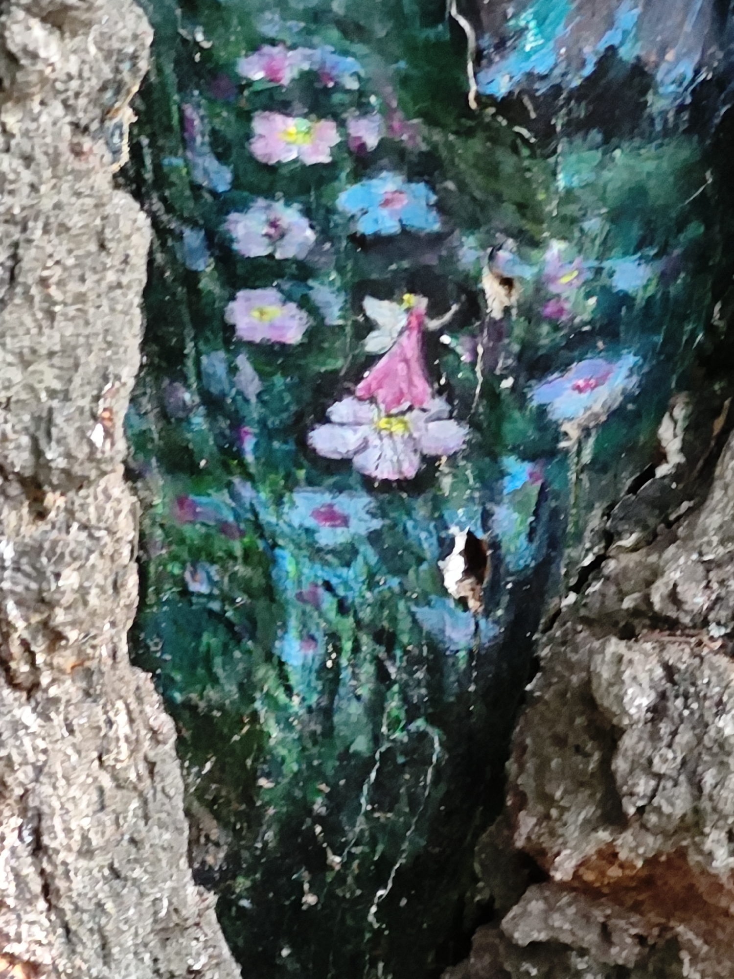 An overhead, close-up, outdoor shot shows a painting on a tree. The painting depicts a fairy in the center, wearing a pink dress and white wings. She is surrounded by flowers in shades of pink and blue, against a dark green, mossy background. The tree bark is visible on the left and right sides of the frame, with a rough texture. The painting appears to be located in a park or garden, given the natural setting and the presence of painted flora.
