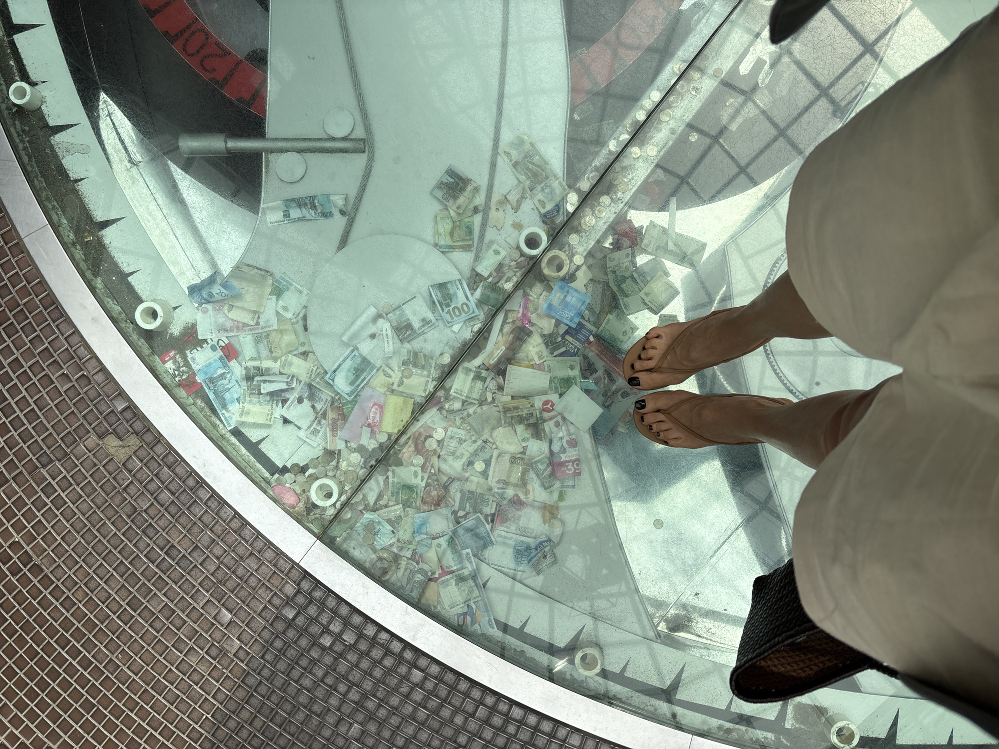 A high-angle shot shows a person standing on a glass floor, wearing sandals and white shorts, in the city of Izhevsk, Russia. Under the glass floor, numerous bills and coins are scattered, possibly as a wishing well or a collection point for monetary donations. The glass floor is circular with geometric shapes. A tiled floor surrounds the glass structure. A person is standing on the floor wearing sandals with black nail polish.