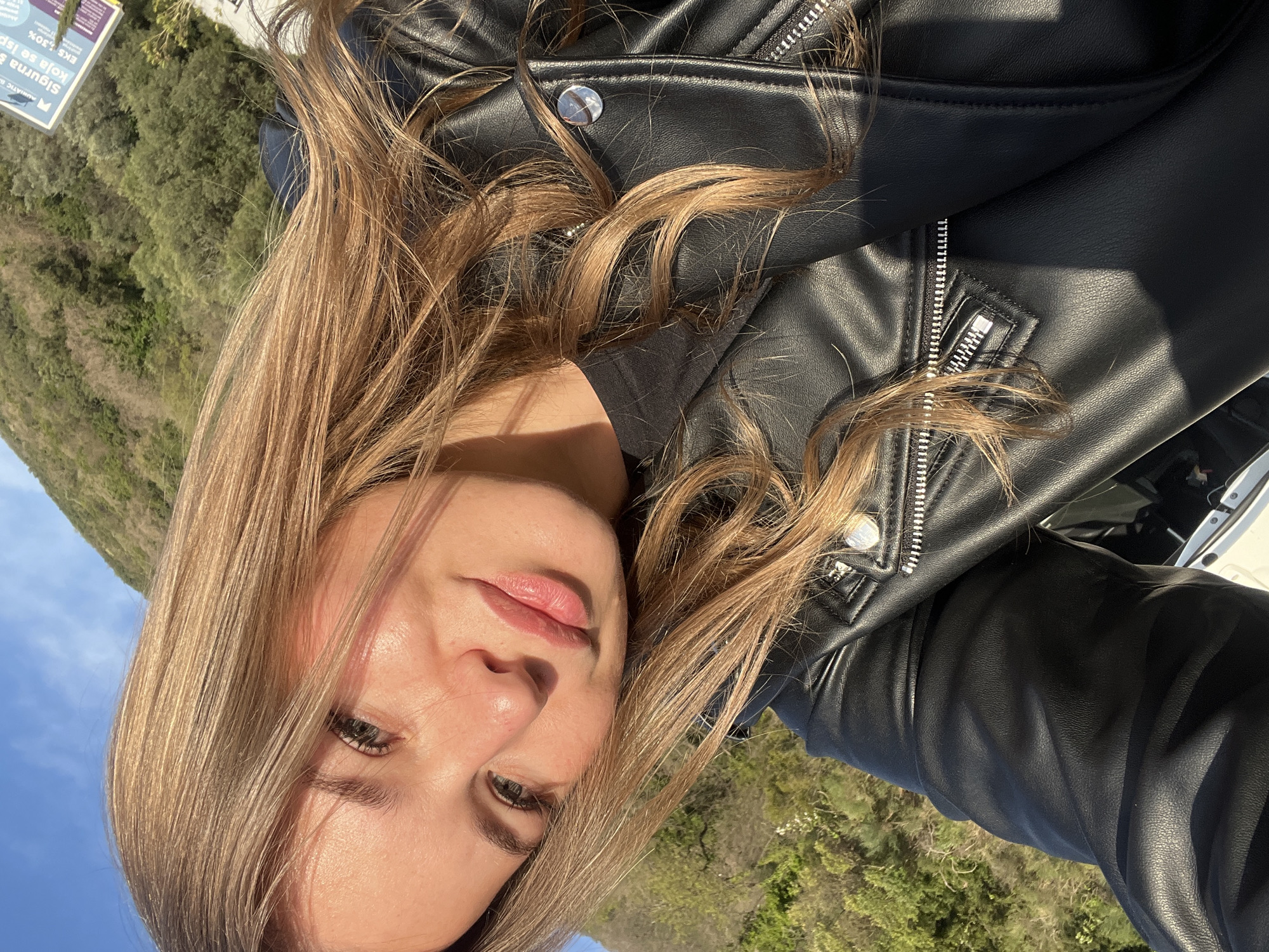 A light-skinned woman with blonde hair is seen in a close-up, rotated headshot, wearing a black leather jacket. The background shows green trees and foliage, along with a blue sky, suggesting an outdoor setting. The location is specified as Prijevor, Montenegro.