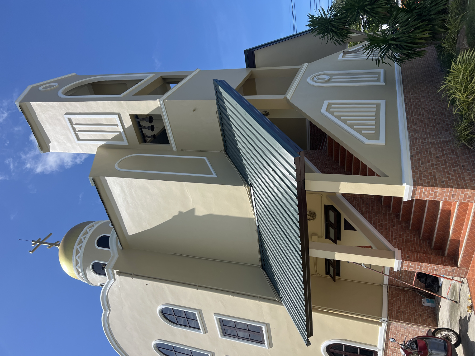 The image shows a tan-colored church with a golden dome and a cross on top, situated against a clear blue sky. The church has a bell tower with three bells visible. There are arched windows on the main building and a green sloped roof over an outdoor staircase leading to the entrance. The staircase is constructed with red-brown brickwork at the base. A red scooter is parked to the left of the stairs, and various construction or cleaning supplies, including a ladder and black trash bags, are present on the ground. Lush green plants and trees are visible on the right side of the image. The scene appears to be in a tropical or subtropical climate, consistent with the location in Koh Phangan, Thailand.