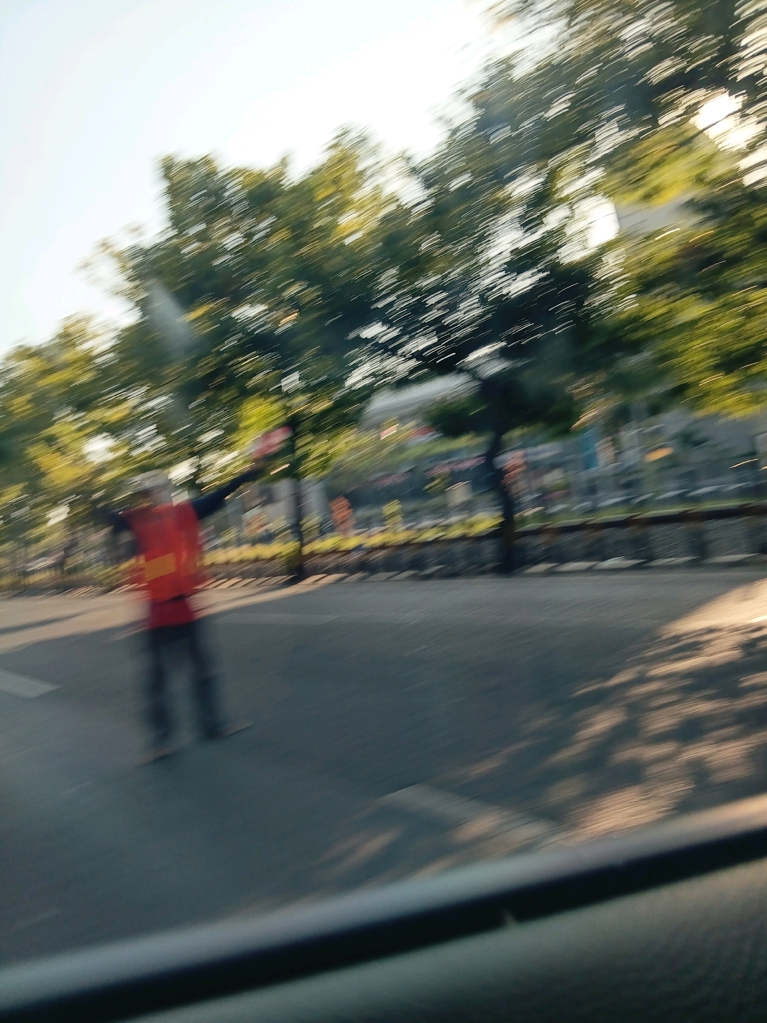 The image depicts a motion-blurred scene of a street in Surabaya, Indonesia, during daylight hours. In the foreground, a person wearing a red vest with yellow accents is visible, with arms outstretched. The person appears to be standing on an asphalt road. Trees with green foliage line the background, and sunlight filters through them, creating dappled shadows on the road. Structures, possibly buildings or traffic infrastructure, are faintly discernible behind the trees. The overall impression is of movement, possibly from a vehicle passing by, as indicated by the streaking effect on the trees and the sky.