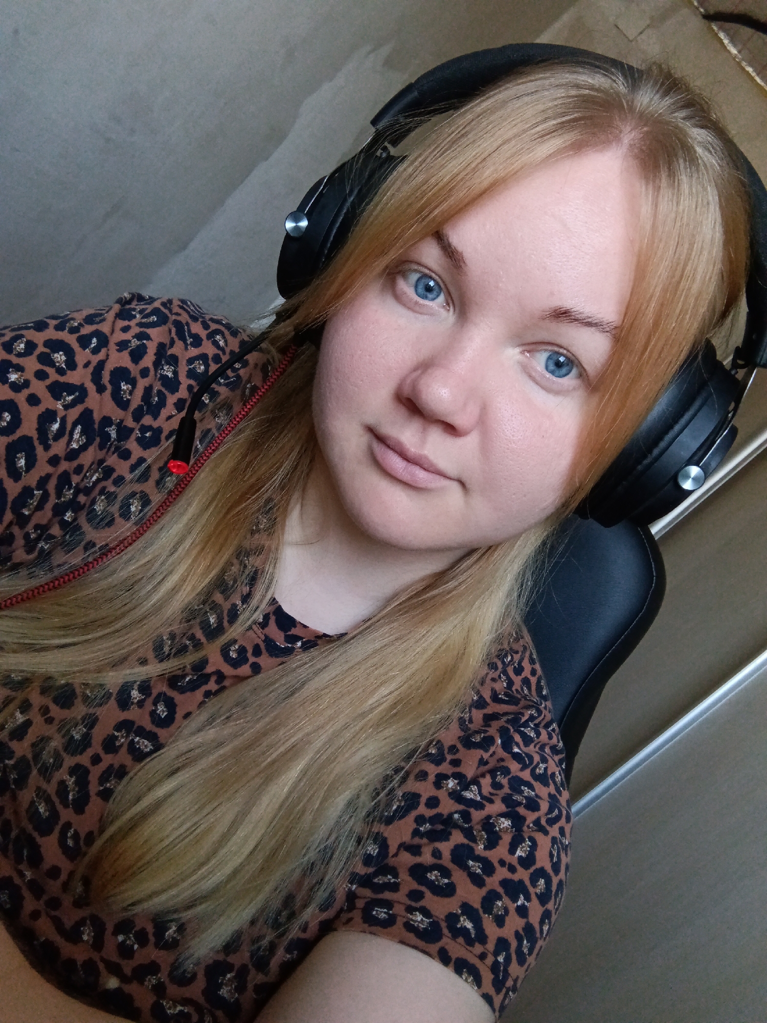 A woman with light blonde hair and blue eyes is wearing black headphones and a brown leopard print shirt. She is sitting in a black chair indoors. The background consists of a beige wall and a silver vertical bar, possibly part of a door frame or shelving unit. The city Фоки, Russia is not discernable in the image.