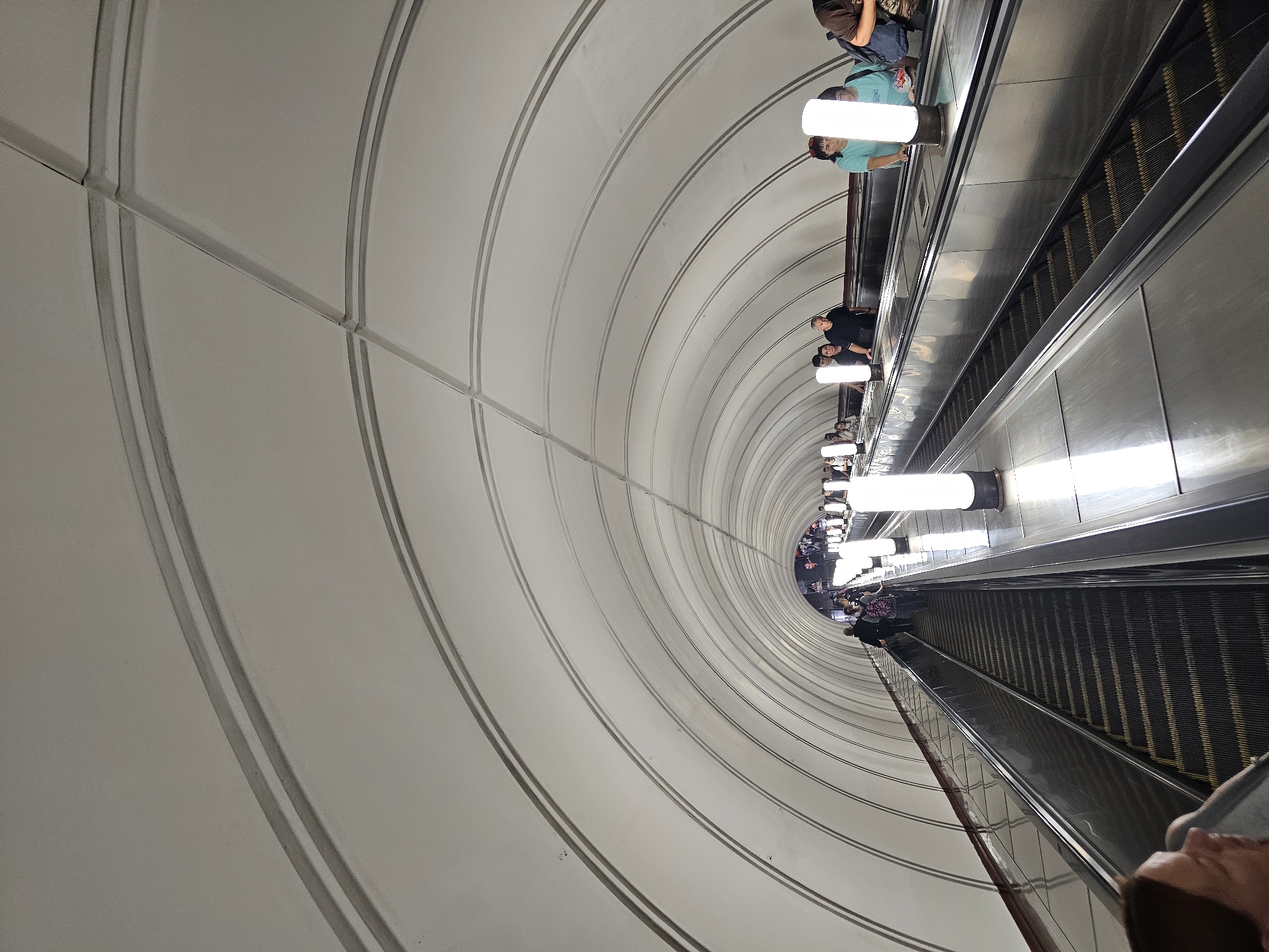 The image captures a scene within the Moscow Metro system, specifically on a long escalator. The escalator, a prominent feature of the image, travels downwards into a vast, arched tunnel. The ceiling of the tunnel is a light, neutral color, with distinctive parallel, curved lines that create a sense of depth and scale. Along the sides of the escalator, vertical, cylindrical light fixtures illuminate the space with a soft, white glow.
Several individuals are present on the escalators, some ascending and others descending. They appear to be everyday commuters, dressed in casual attire. Their expressions and postures suggest a typical metro experience, with some individuals looking straight ahead, while others may be engaged in conversation or looking at their phones. The escalators themselves are metallic, with visible steps and handrails. The overall atmosphere is one of public transportation in use, with a sense of movement and activity. The lighting and architectural style are characteristic of many Moscow Metro stations, known for their grandeur and unique designs.
