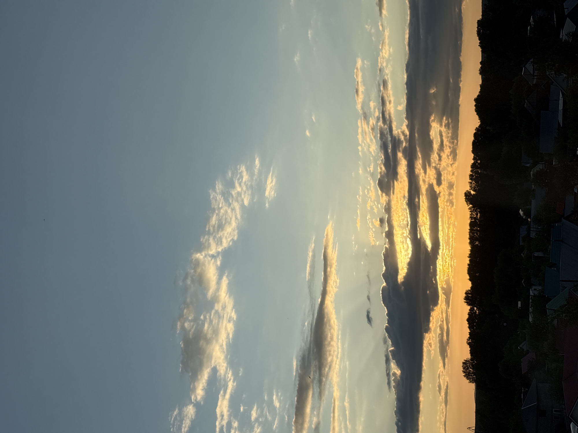 An evening sky over Chaikovsky, Russia, displays a sunset with warm hues of orange and yellow peeking through scattered clouds. The horizon is marked by a dark silhouette of trees, behind which a few buildings with colorful roofs are faintly visible. The sky transitions from a bright orange near the horizon to a soft blue overhead.