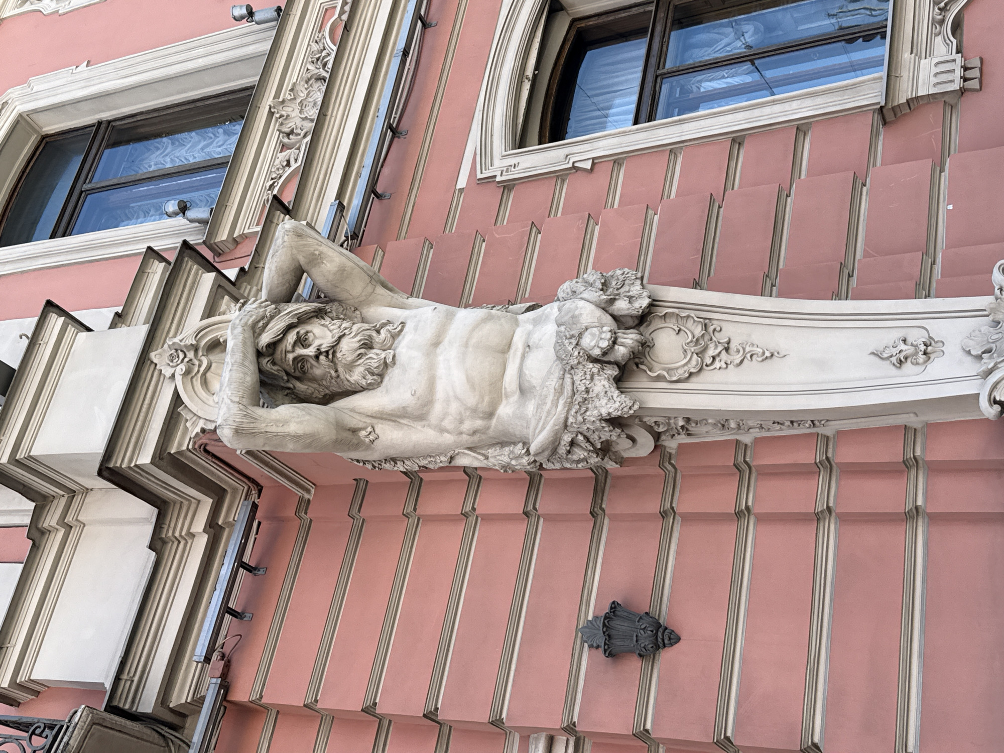 This media file shows a low-angle, close-up, outdoor view of a building facade in Saint Petersburg, Russia. The building is adorned with elaborate architectural details, including sculpted ornamentation and decorative elements. The primary subject is a large, white marble sculpture of a reclining male figure, likely a mythological or allegorical representation, positioned prominently on the facade. The figure has a bearded face, muscular physique, and is depicted with arms raised behind his head. The sculpture is integrated with the building's decorative framework, which features ornate carvings and moldings.
The building itself is painted a soft pink color and features a series of recessed panels with light-colored trim, creating a sense of depth and texture. Above the sculpture, there are windows with dark frames and reflective glass, hinting at the interior or sky. The architectural style suggests a historical or classical influence, common in Saint Petersburg.
There is no discernible activity or event taking place; the scene is static, focusing on the architectural artistry. Notable details include the intricate sculpting of the figure and the ornamental details around it. The lighting suggests it is daytime, but the weather is not apparent. The overall impression is one of grandeur and artistic craftsmanship, characteristic of the city's historical architecture. No visible text is present in the image.