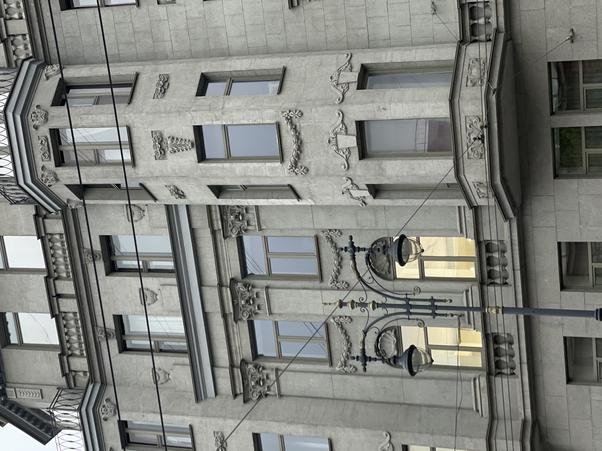 The image depicts a building in Saint Petersburg, Russia. The building has a gray facade with rectangular windows. The windows have decorative elements and mouldings around them. A street lamp with two spherical lamps is attached to the building's facade, partially obscuring the view of some windows. Overhead electrical wires run diagonally across the image, and other buildings are visible in the background.