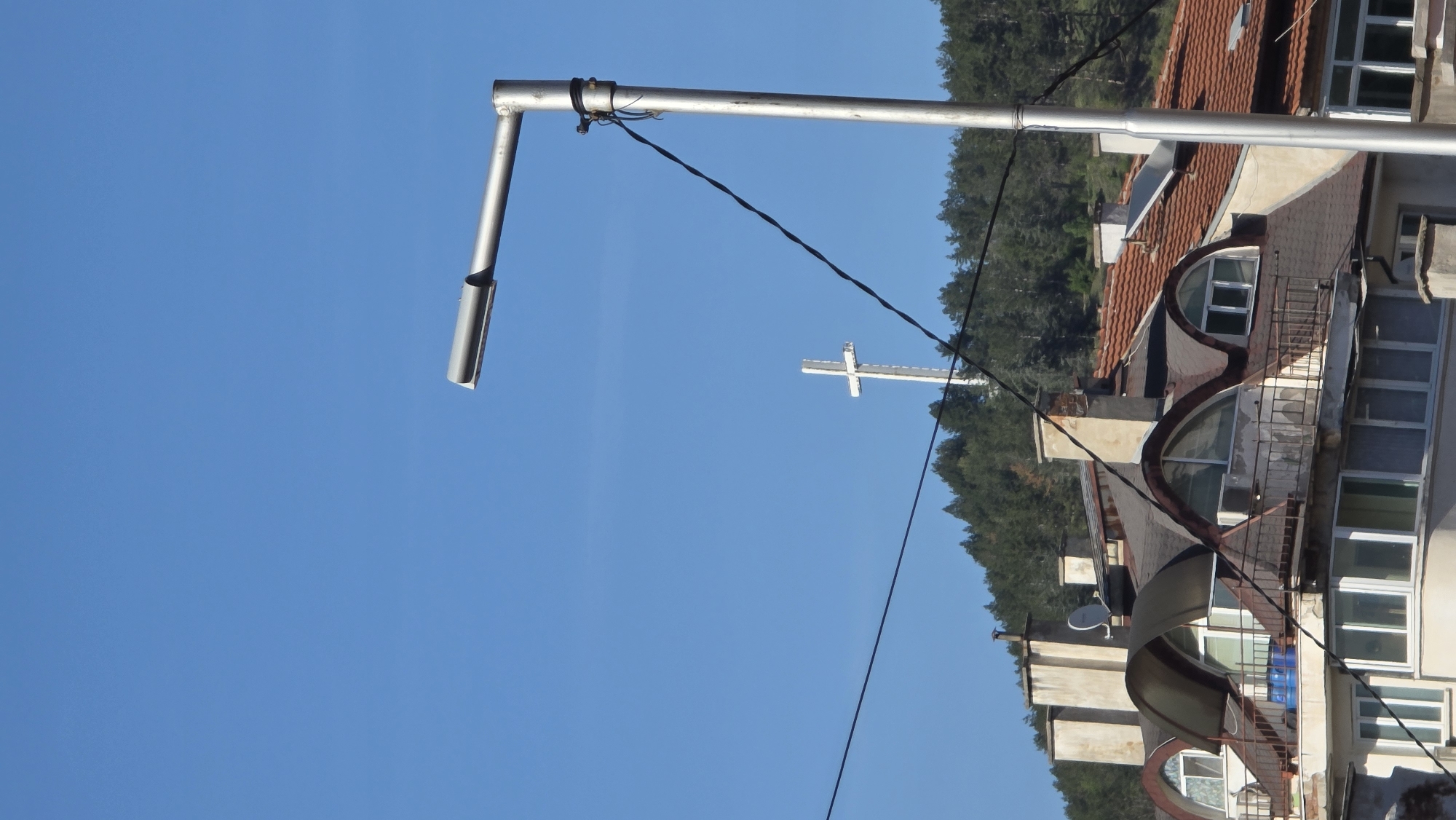 The image shows a daytime view of a building and surrounding elements in Blagoevgrad, Bulgaria. The sky is clear and blue. In the foreground, a silver metal streetlight pole is visible. An electrical wire stretches across the sky, connecting to the pole. In the background, a large, white cross sits atop a hill covered with trees. To the right, part of a building is visible, with a red tile roof and balconies. Some of the building windows and architectural features are evident. The building is situated amidst trees.