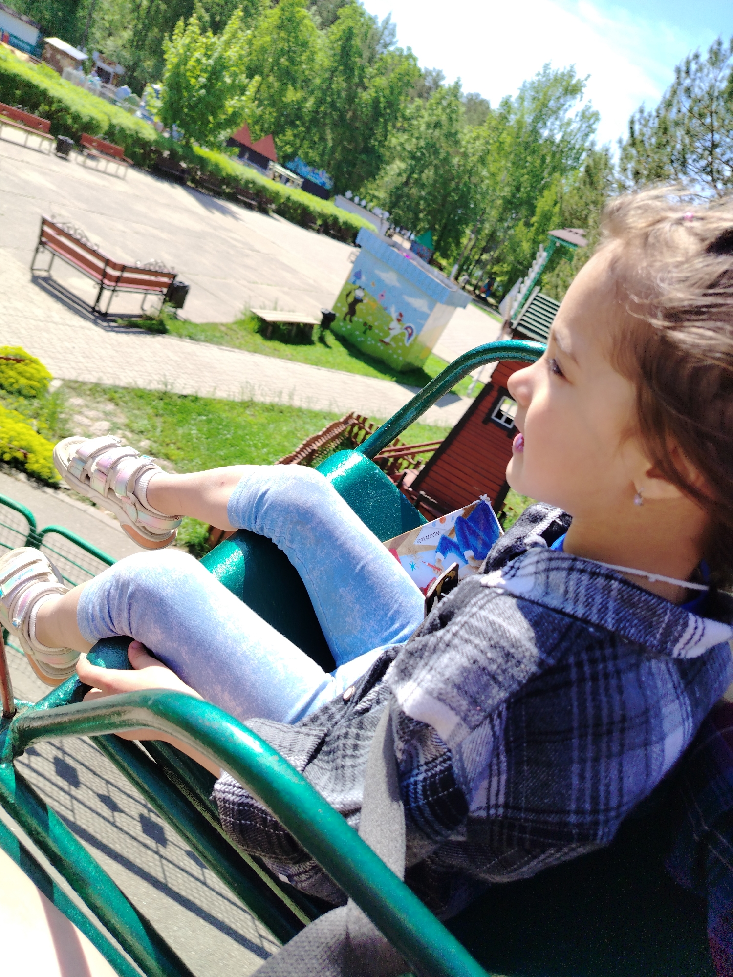 A girl rides an amusement park ride on a sunny day. She wears blue leggings, pink sandals, and a gray plaid shirt over a darker top. The ride appears to be a small spinning carousel, with the girl seated in a green seat. In the background, there is a park with benches, trees, and a small building. The location is Chaikovsky, Russia, based on landmarks.