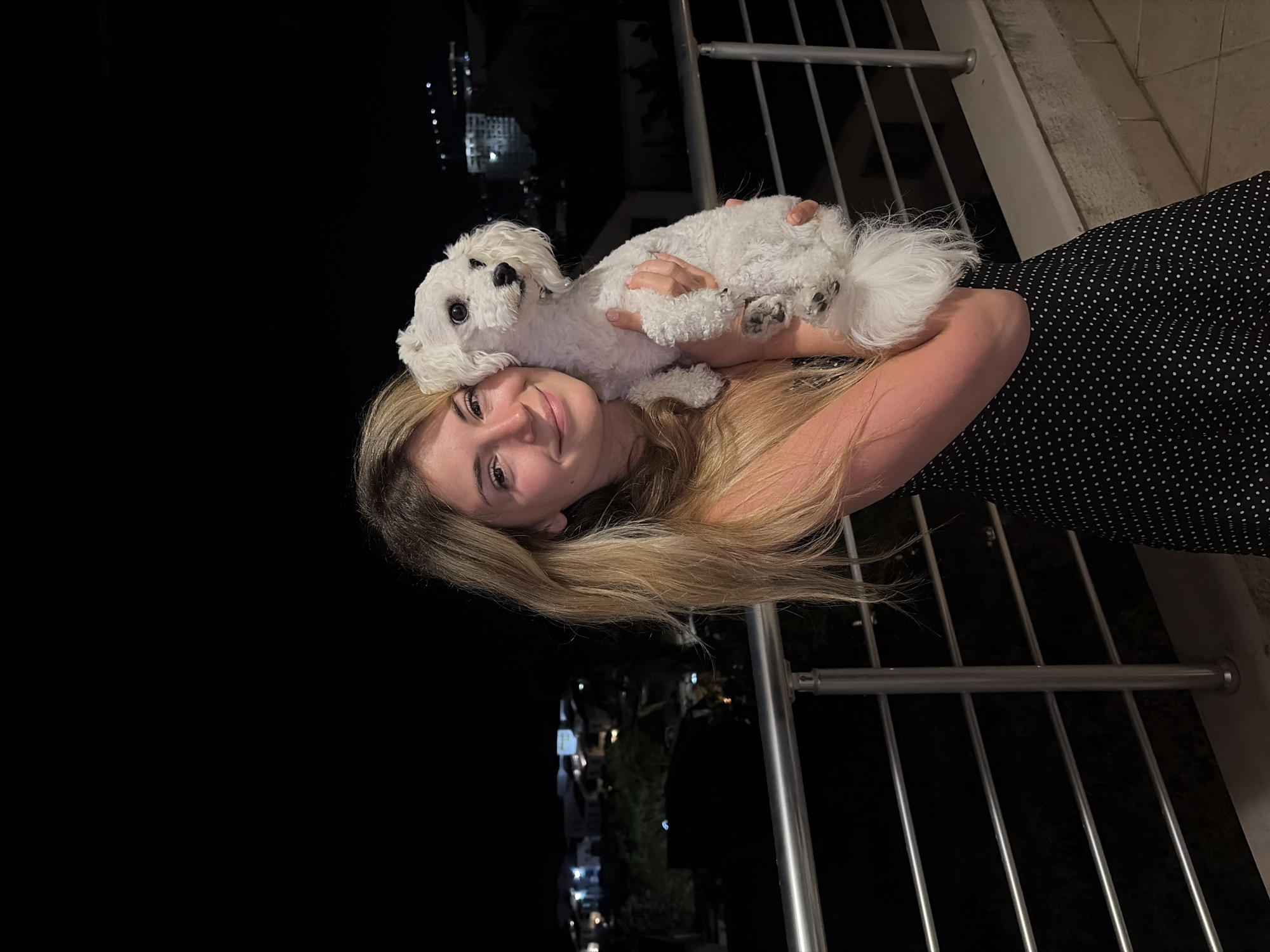 A young woman with long blonde hair is holding a small white dog in her arms, likely a Maltese or similar breed. She is smiling slightly at the camera. The background appears to be a nighttime scene, potentially a balcony or outdoor area. The presence of a metal railing suggests she might be on a balcony or elevated space. In the dark background, one can see scattered lights, indicating a city environment. It is possible the picture was taken in the city of Budva, Montenegro due to the mention of the place. The woman is wearing a black dress with white polka dots.