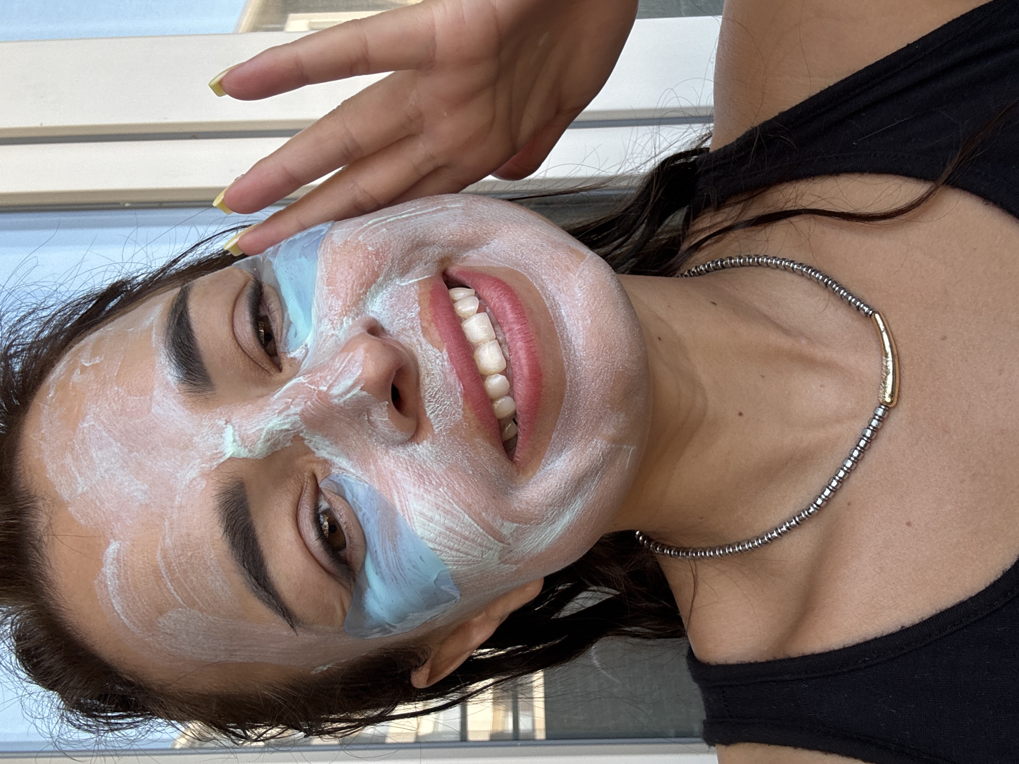 A medium shot, close-up of a woman's face as she applies a face mask. She is smiling with her mouth open, revealing white teeth. She has brown eyes, dark eyebrows, and dark hair pulled back. Her face is covered in a white face mask and light blue under-eye patches. Her right hand is raised and touching her temple. She is wearing a black tank top and a silver necklace with a gold pendant. The background is blurred and appears to be outdoors with a window or building visible. The lighting suggests it is daytime.